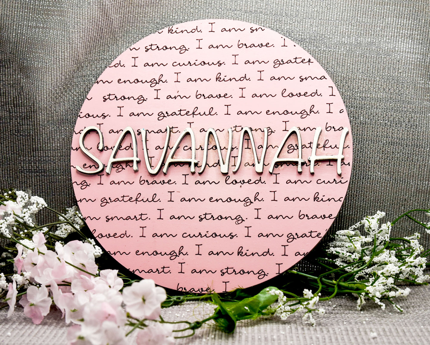 Round wooden name affirmation sign with a custom 3D name on top and engraved positive words like "brave," "kind," and "loved" behind it. Offered in various colors and three sizes, it's perfect for crib name tags, baby announcements, or personalized nursery decor.