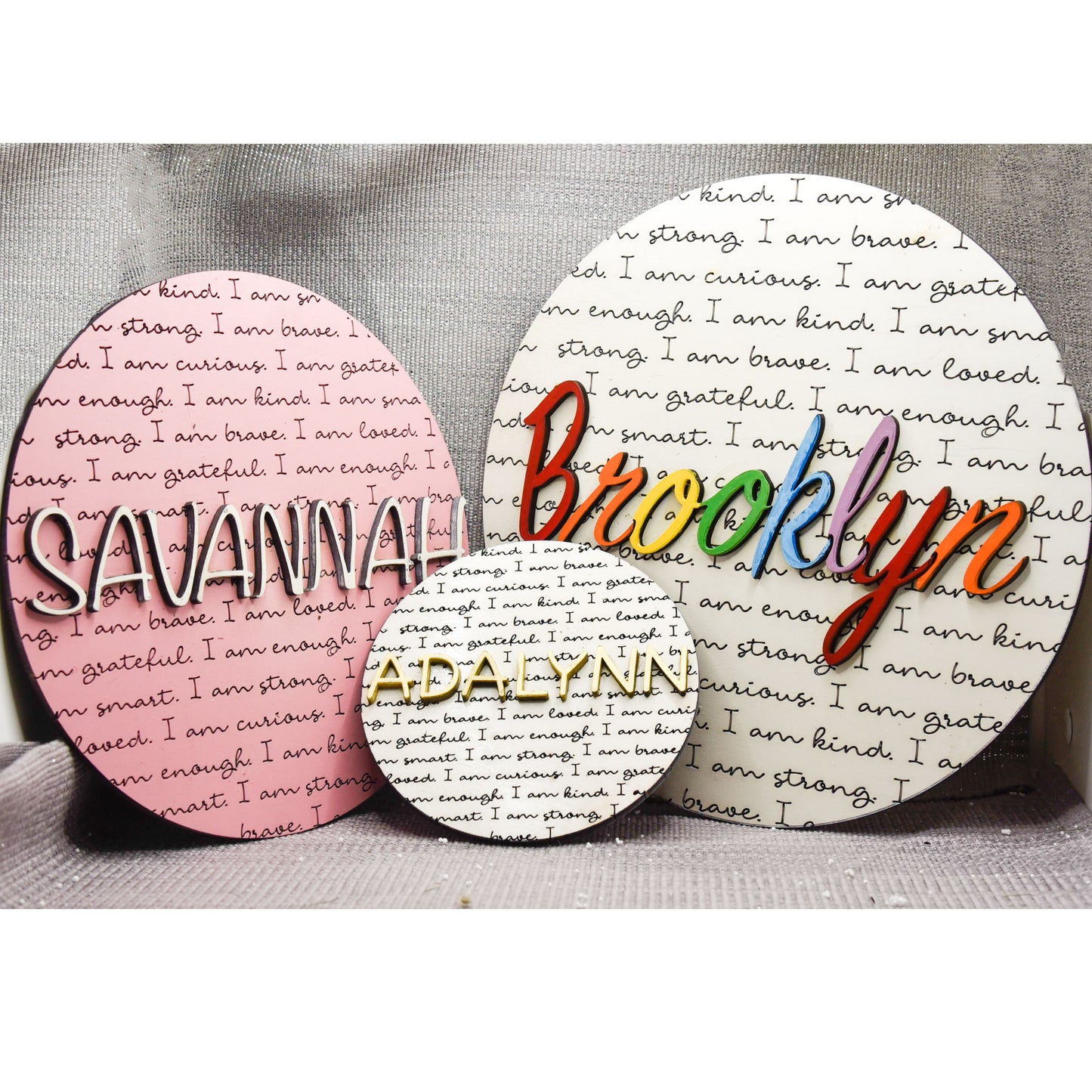 Round wooden name affirmation sign with a custom 3D name on top and engraved positive words like "brave," "kind," and "loved" behind it. Offered in various colors and three sizes, it's perfect for crib name tags, baby announcements, or personalized nursery decor.