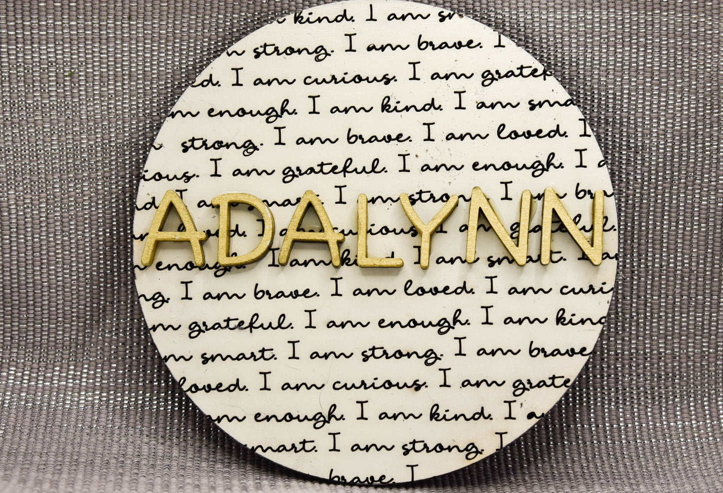 Round wooden name affirmation sign with a custom 3D name on top and engraved positive words like "brave," "kind," and "loved" behind it. Offered in various colors and three sizes, it's perfect for crib name tags, baby announcements, or personalized nursery decor.