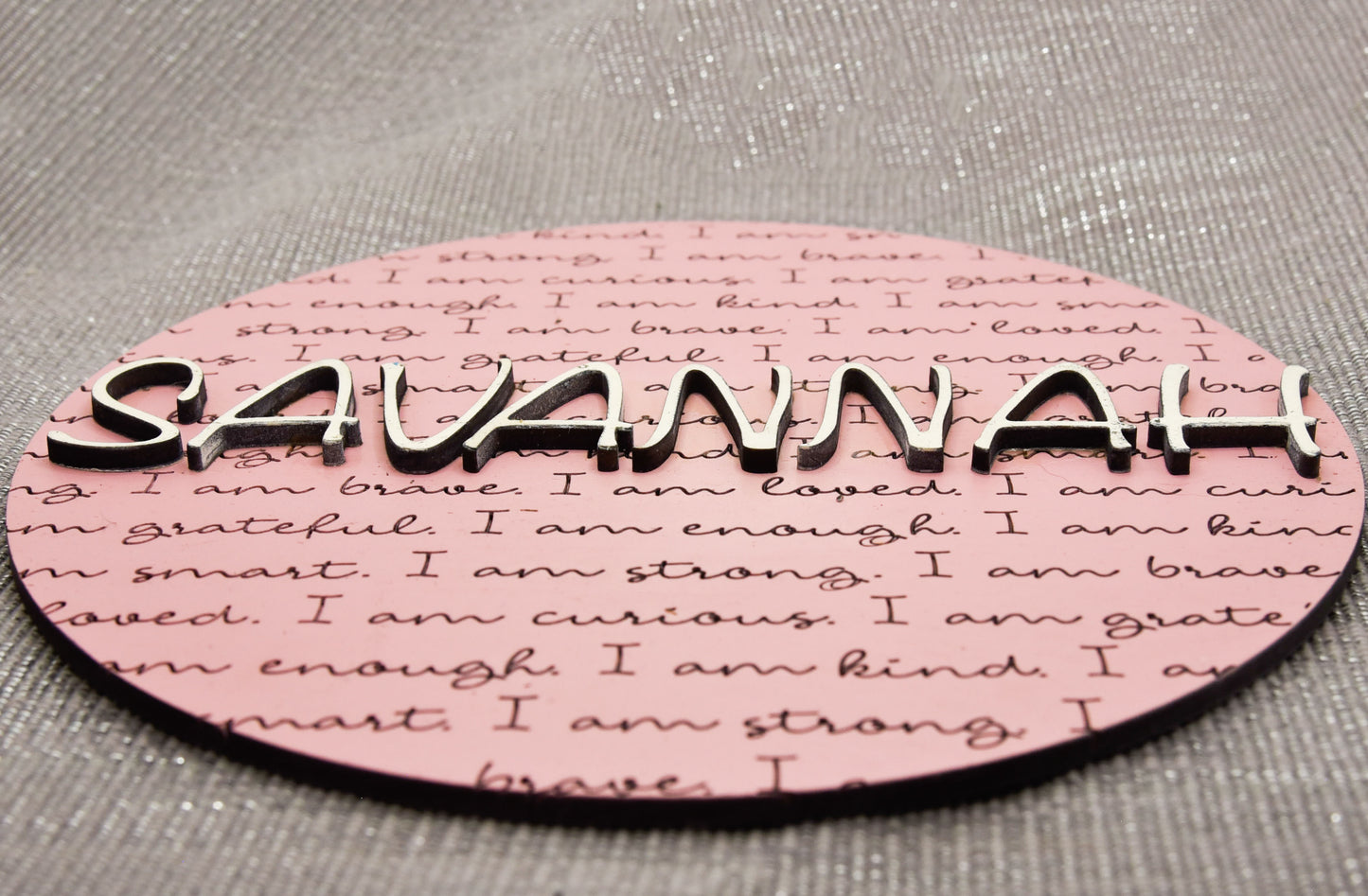 Round wooden name affirmation sign with a custom 3D name on top and engraved positive words like "brave," "kind," and "loved" behind it. Offered in various colors and three sizes, it's perfect for crib name tags, baby announcements, or personalized nursery decor.