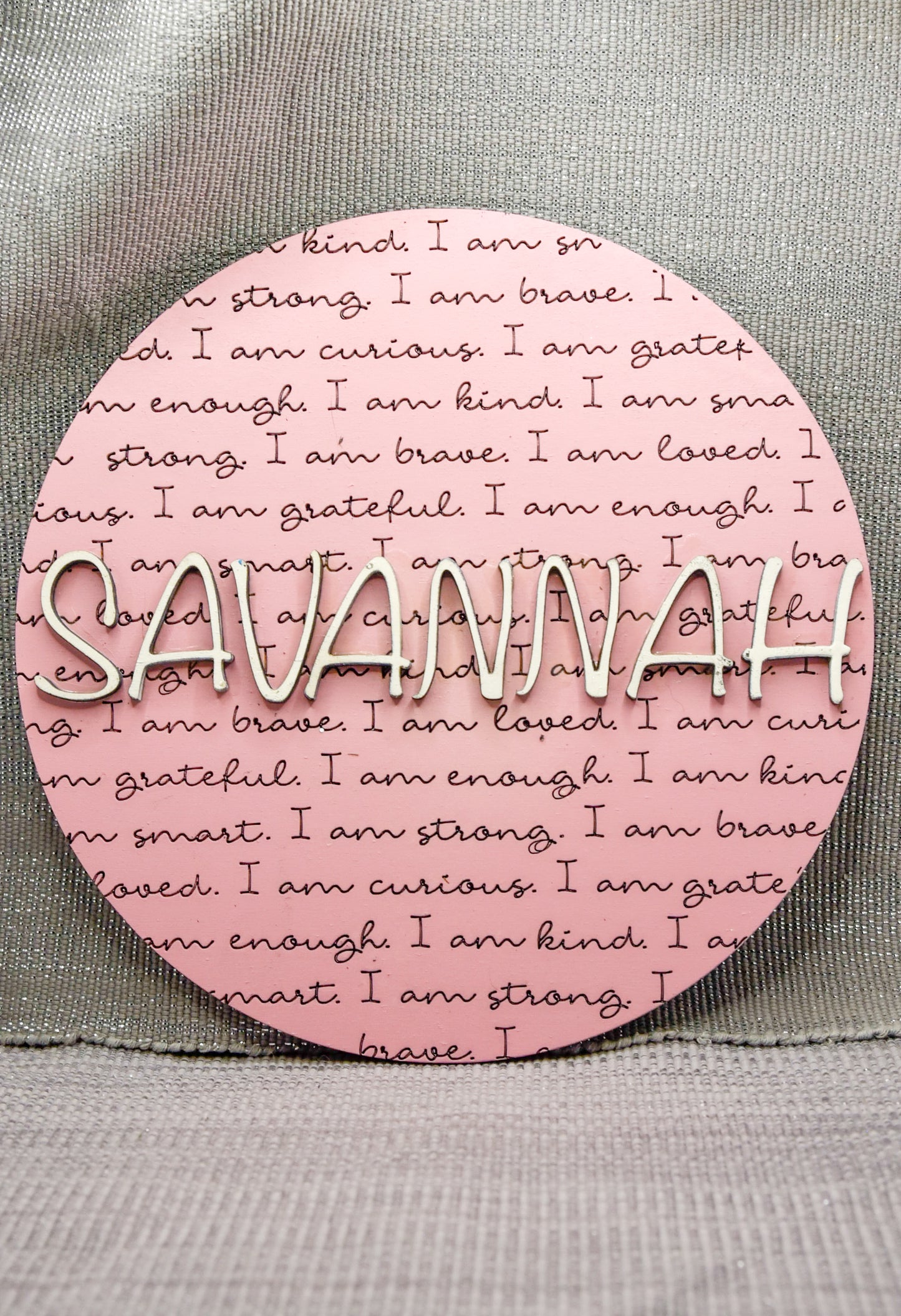 Round wooden name affirmation sign with a custom 3D name on top and engraved positive words like "brave," "kind," and "loved" behind it. Offered in various colors and three sizes, it's perfect for crib name tags, baby announcements, or personalized nursery decor.
