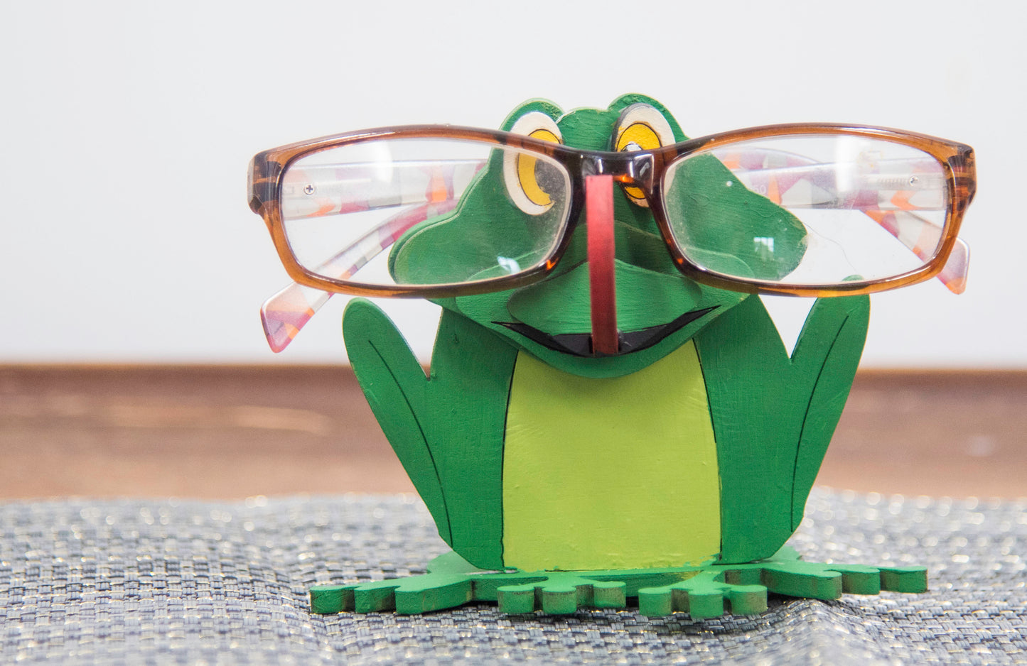 Hand-painted wooden frog-shaped glasses holder with bulging eyes and a flat base to hold eyeglasses. Ideal for desks, nightstands, or gifting to frog lovers and animal fans