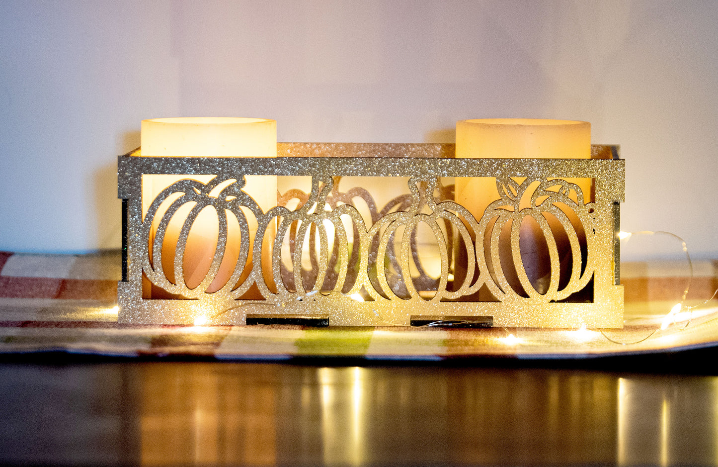 et of three intricately designed metal and wood pumpkin candle holders displayed on a checkered fall table runner. The set includes a tall rectangular lantern, a horizontal centerpiece tray, and a small square holder, each featuring layered pumpkin cutouts that beautifully glow with the warm light of tea candles inside—perfect for cozy fall, Thanksgiving, or farmhouse-style decor.