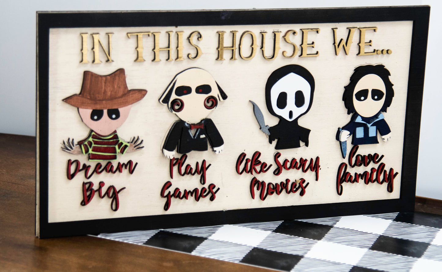 Wooden Halloween sign featuring layered cutout horror movie characters with engraved phrase “In This House,” handcrafted and freestanding, designed for spooky home decor fans.