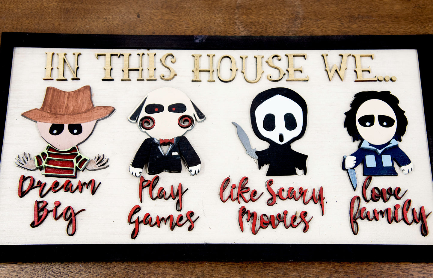 Wooden Halloween sign featuring layered cutout horror movie characters with engraved phrase “In This House,” handcrafted and freestanding, designed for spooky home decor fans.