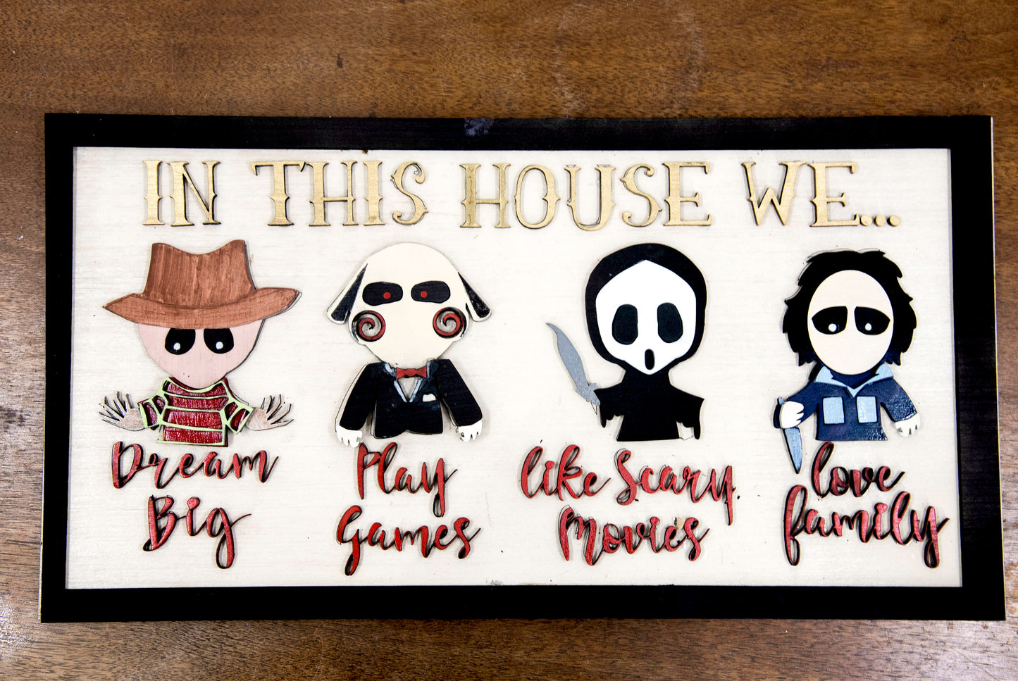 Wooden Halloween sign featuring layered cutout horror movie characters with engraved phrase “In This House,” handcrafted and freestanding, designed for spooky home decor fans.
