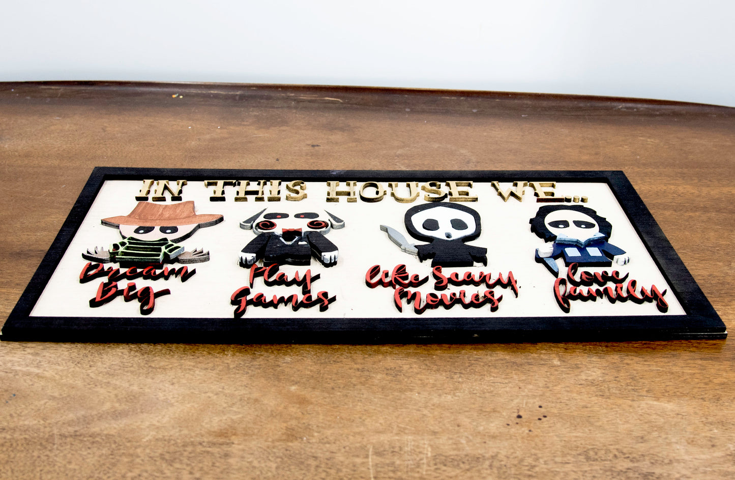 Wooden Halloween sign featuring layered cutout horror movie characters with engraved phrase “In This House,” handcrafted and freestanding, designed for spooky home decor fans.