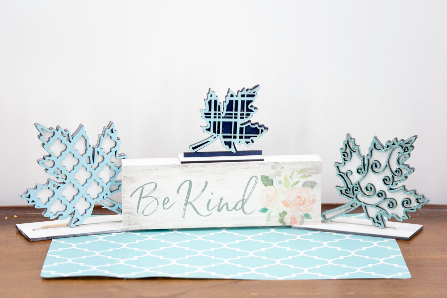 Handcrafted standing wooden leaf decor pieces in layered colors and patterns. Each leaf has a stained background with a cutout patterned top layer, sitting on a flat wood base. Ideal for fall or farmhouse-style decor