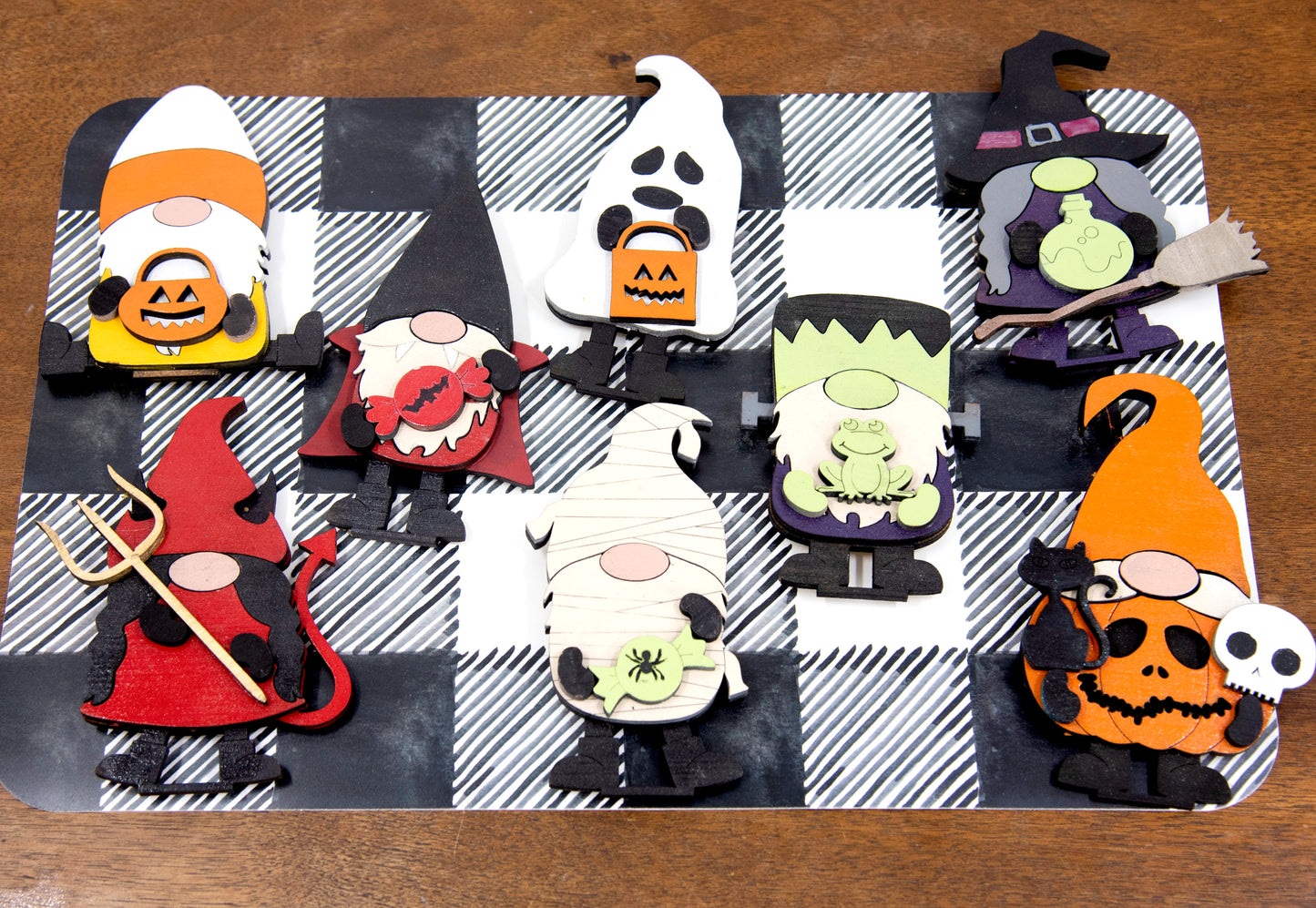 Set of hand-painted wooden Halloween gnome figurines dressed in spooky costumes including witch, ghost, pumpkin, vampire, and Frankenstein, standing on a shelf with black and white buffalo check runner.