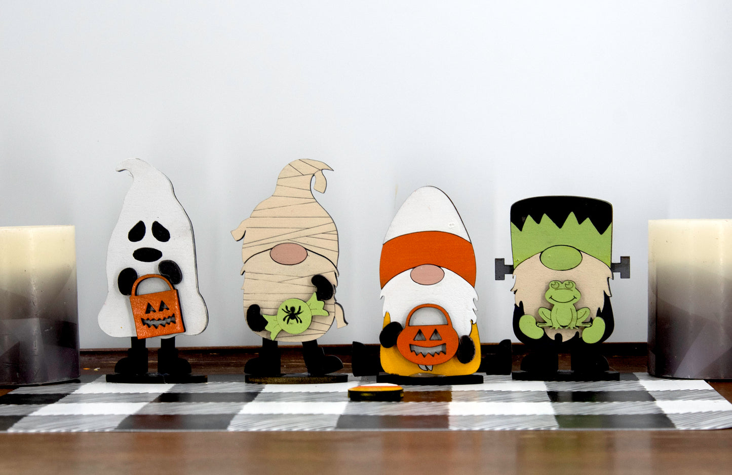 Set of hand-painted wooden Halloween gnome figurines dressed in spooky costumes including witch, ghost, pumpkin, vampire, and Frankenstein, standing on a shelf with black and white buffalo check runner.