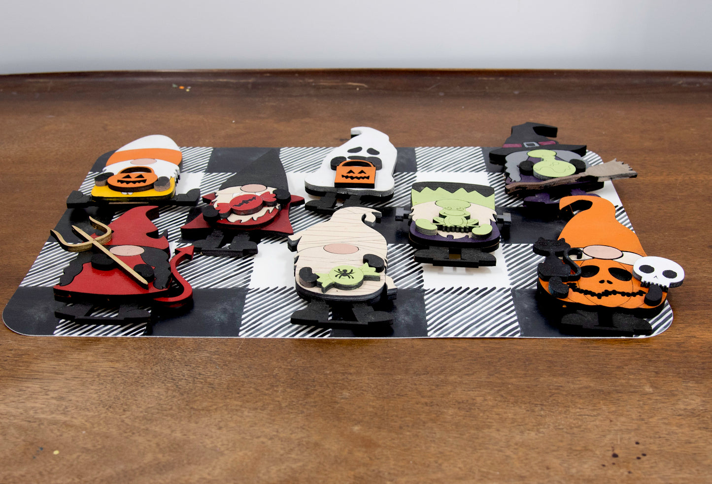 Set of hand-painted wooden Halloween gnome figurines dressed in spooky costumes including witch, ghost, pumpkin, vampire, and Frankenstein, standing on a shelf with black and white buffalo check runner.