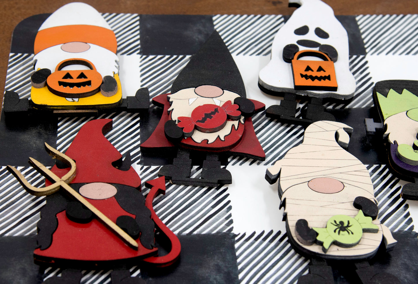 Set of hand-painted wooden Halloween gnome figurines dressed in spooky costumes including witch, ghost, pumpkin, vampire, and Frankenstein, standing on a shelf with black and white buffalo check runner.