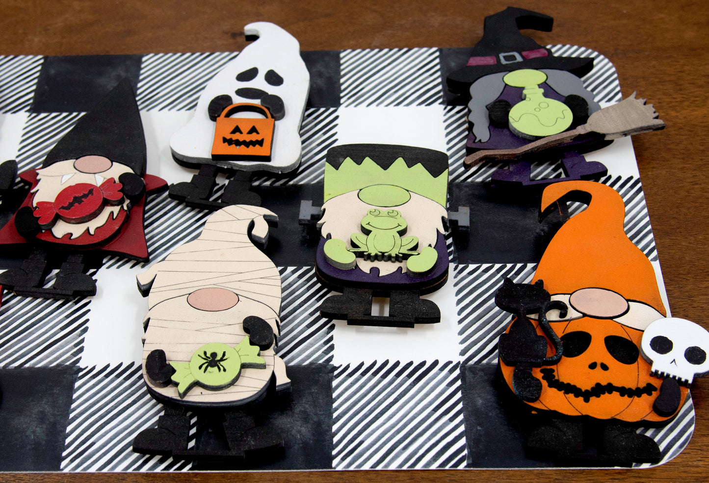 Set of hand-painted wooden Halloween gnome figurines dressed in spooky costumes including witch, ghost, pumpkin, vampire, and Frankenstein, standing on a shelf with black and white buffalo check runner.