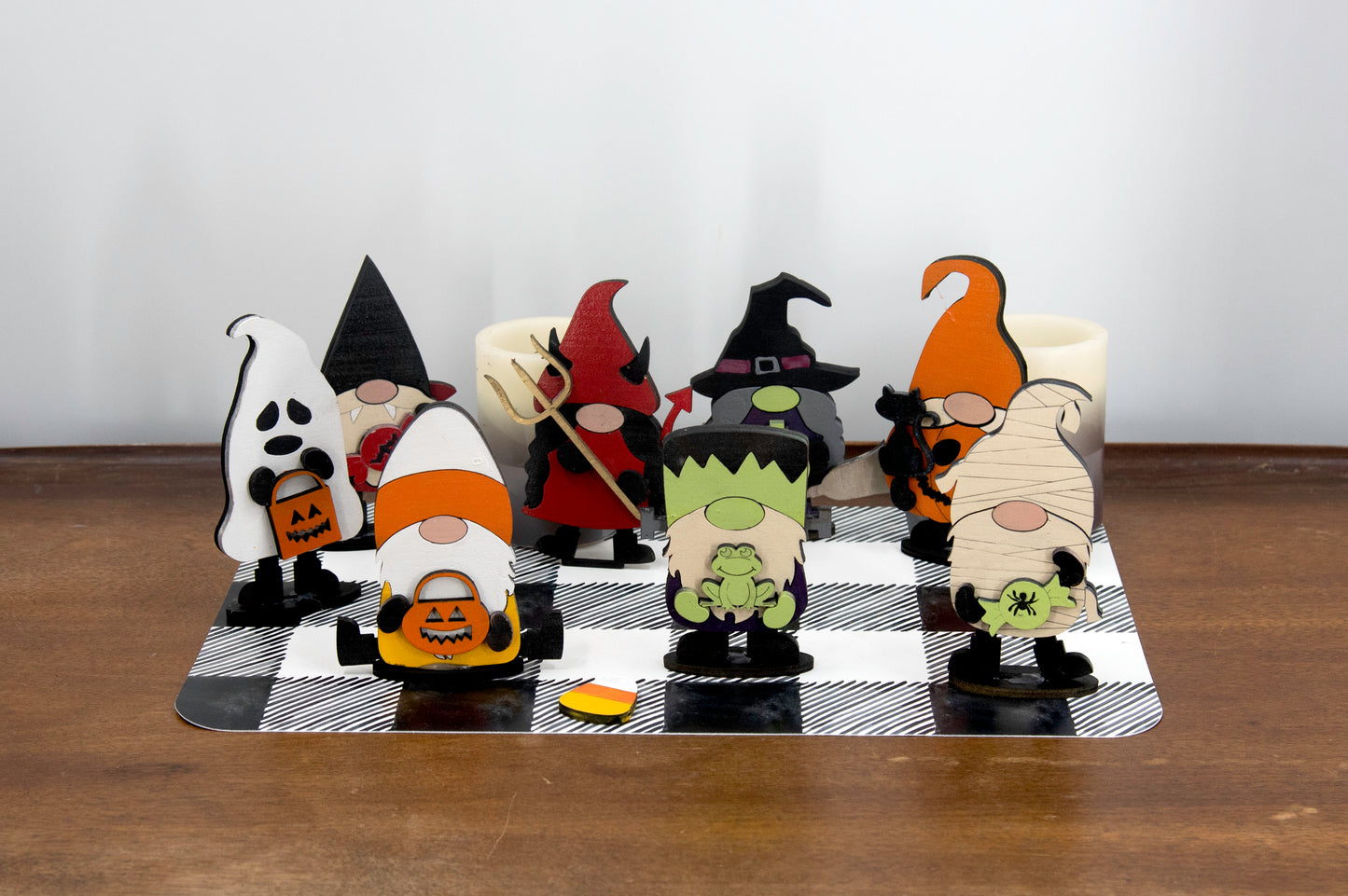 Set of hand-painted wooden Halloween gnome figurines dressed in spooky costumes including witch, ghost, pumpkin, vampire, and Frankenstein, standing on a shelf with black and white buffalo check runner.