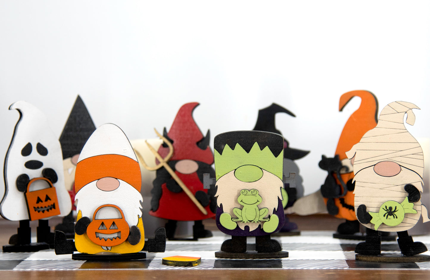 Set of hand-painted wooden Halloween gnome figurines dressed in spooky costumes including witch, ghost, pumpkin, vampire, and Frankenstein, standing on a shelf with black and white buffalo check runner.
