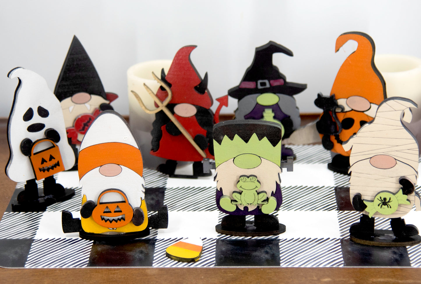 Set of hand-painted wooden Halloween gnome figurines dressed in spooky costumes including witch, ghost, pumpkin, vampire, and Frankenstein, standing on a shelf with black and white buffalo check runner.