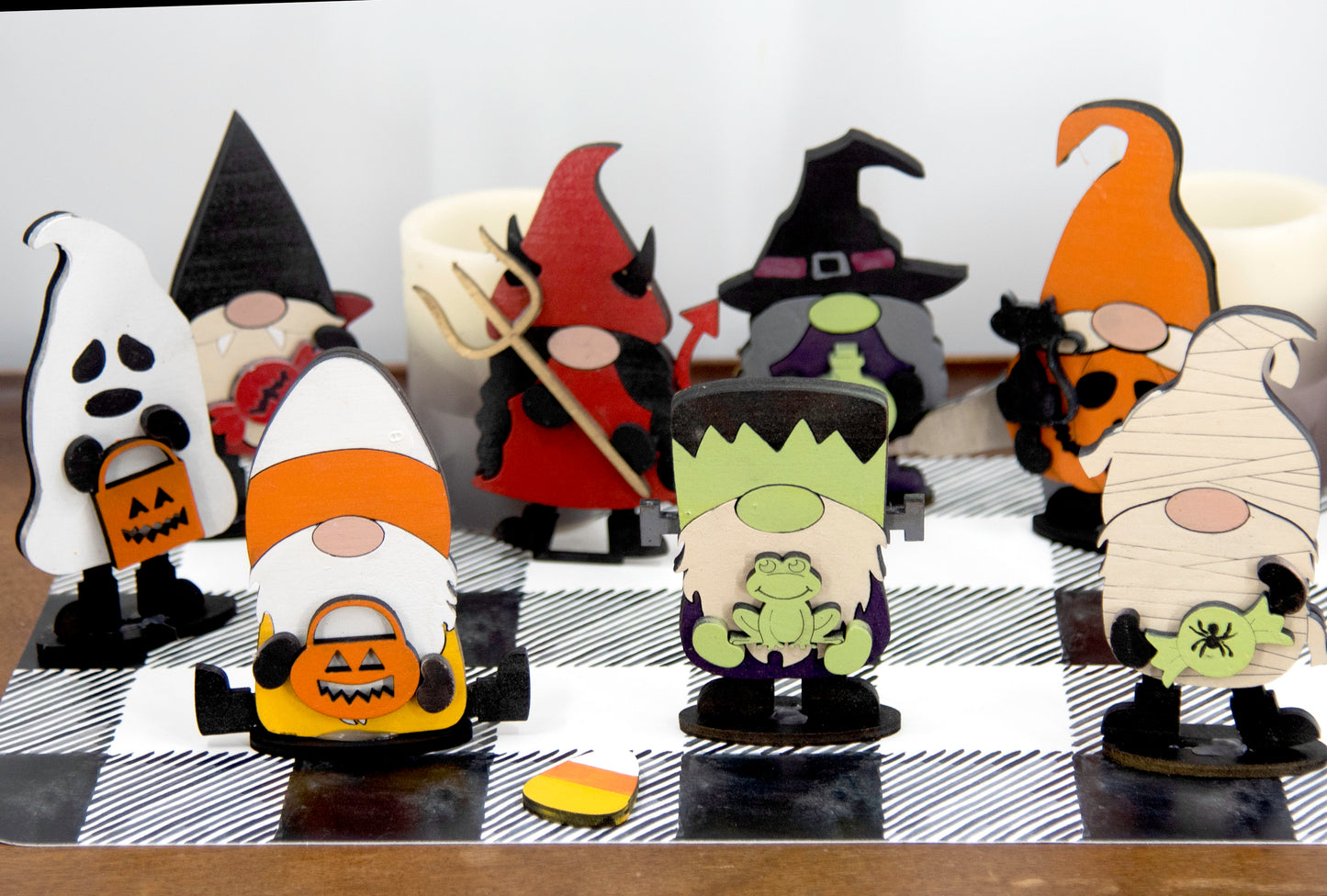 Set of hand-painted wooden Halloween gnome figurines dressed in spooky costumes including witch, ghost, pumpkin, vampire, and Frankenstein, standing on a shelf with black and white buffalo check runner.