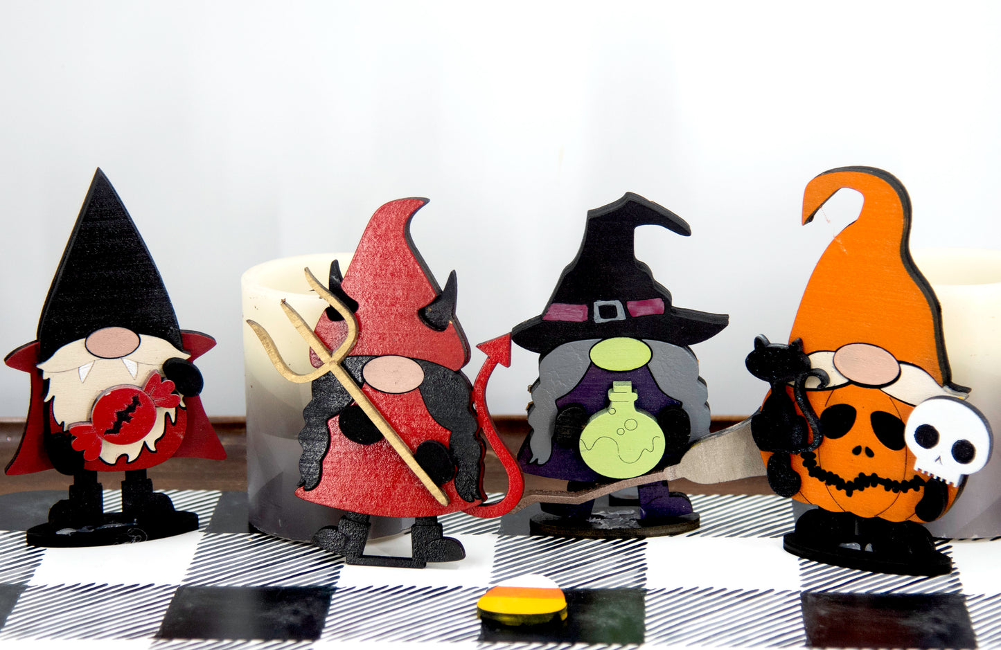 Set of hand-painted wooden Halloween gnome figurines dressed in spooky costumes including witch, ghost, pumpkin, vampire, and Frankenstein, standing on a shelf with black and white buffalo check runner.