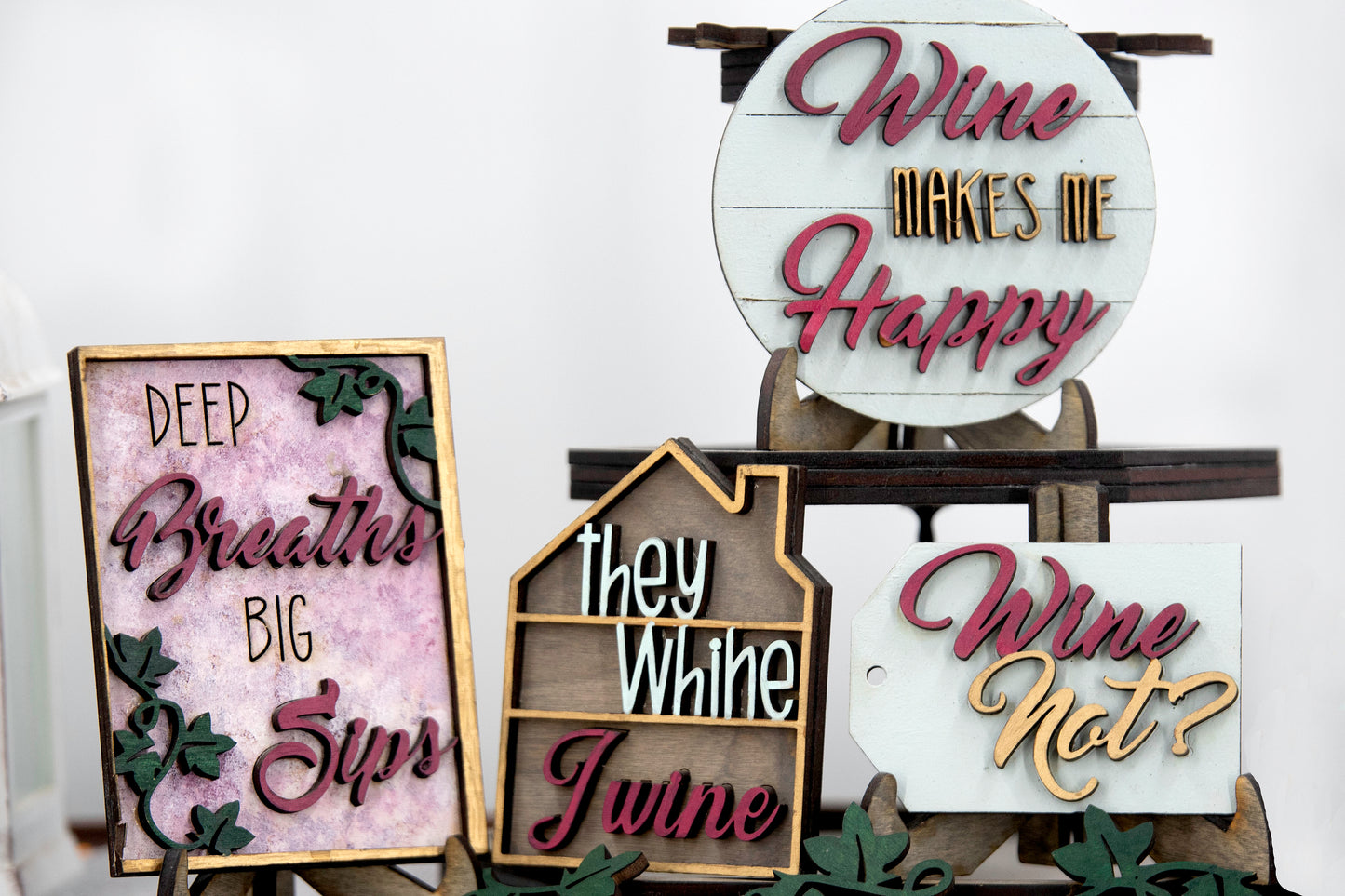 A 6-piece wine-themed wooden tiered tray set featuring humorous mom quotes like "They whine, I wine" and "Mommy juice," painted in maroon, grey, gold, and light grey with included stands and optional tiered tray