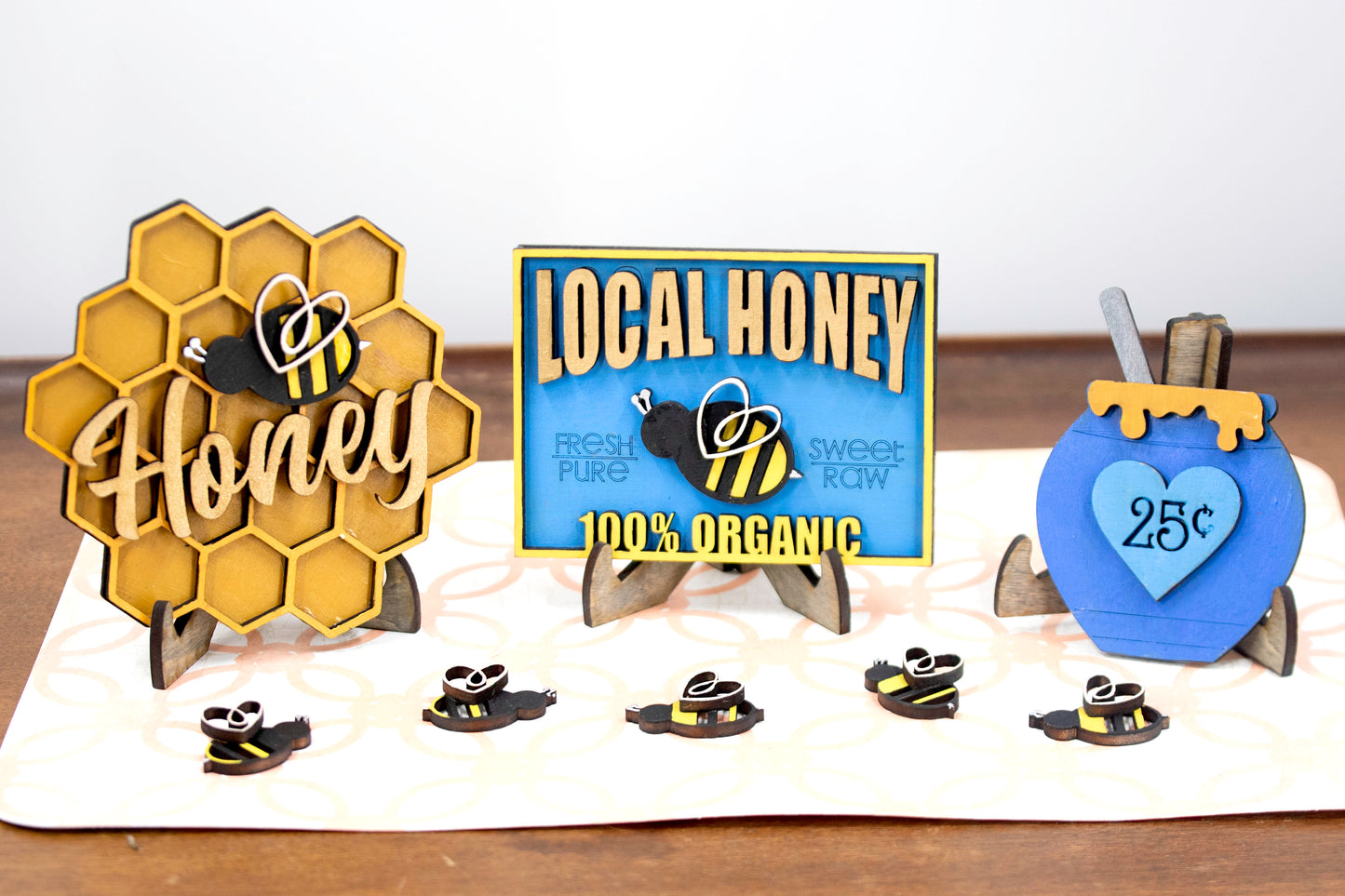DIY bee tiered tray kit with 6 unfinished wood pieces including honey jar, local honey sign, welcome to our hive sign, honey bomb, bee hexagon plaque, and 5 small bee accents. Ideal for painting, staining, and customizing. Comes with stands for easy display. Perfect for summer crafts, farmhouse style decor, or paint-your-own projects. Great for hobby crafters, DIY parties, and seasonal decorating fun with a bee and honey theme.