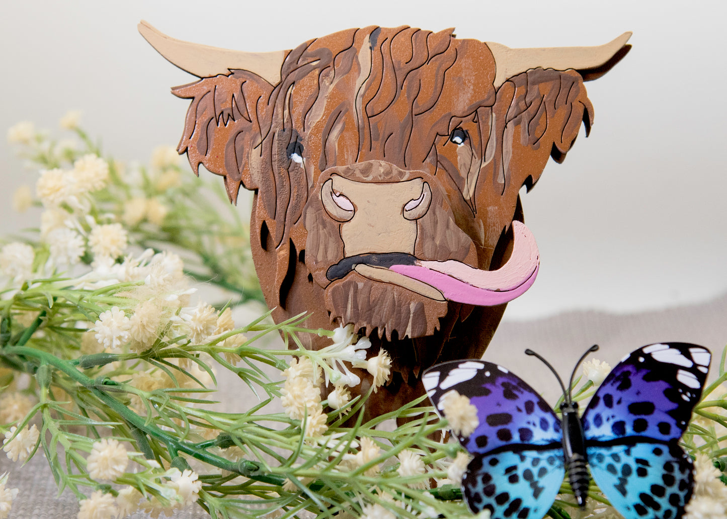Wooden Highland Cow glasses holder with layered fur detail and curled horns, wearing eyeglasses and sticking out its tongue, surrounded by flowers and a decorative butterfly for a rustic and playful look.