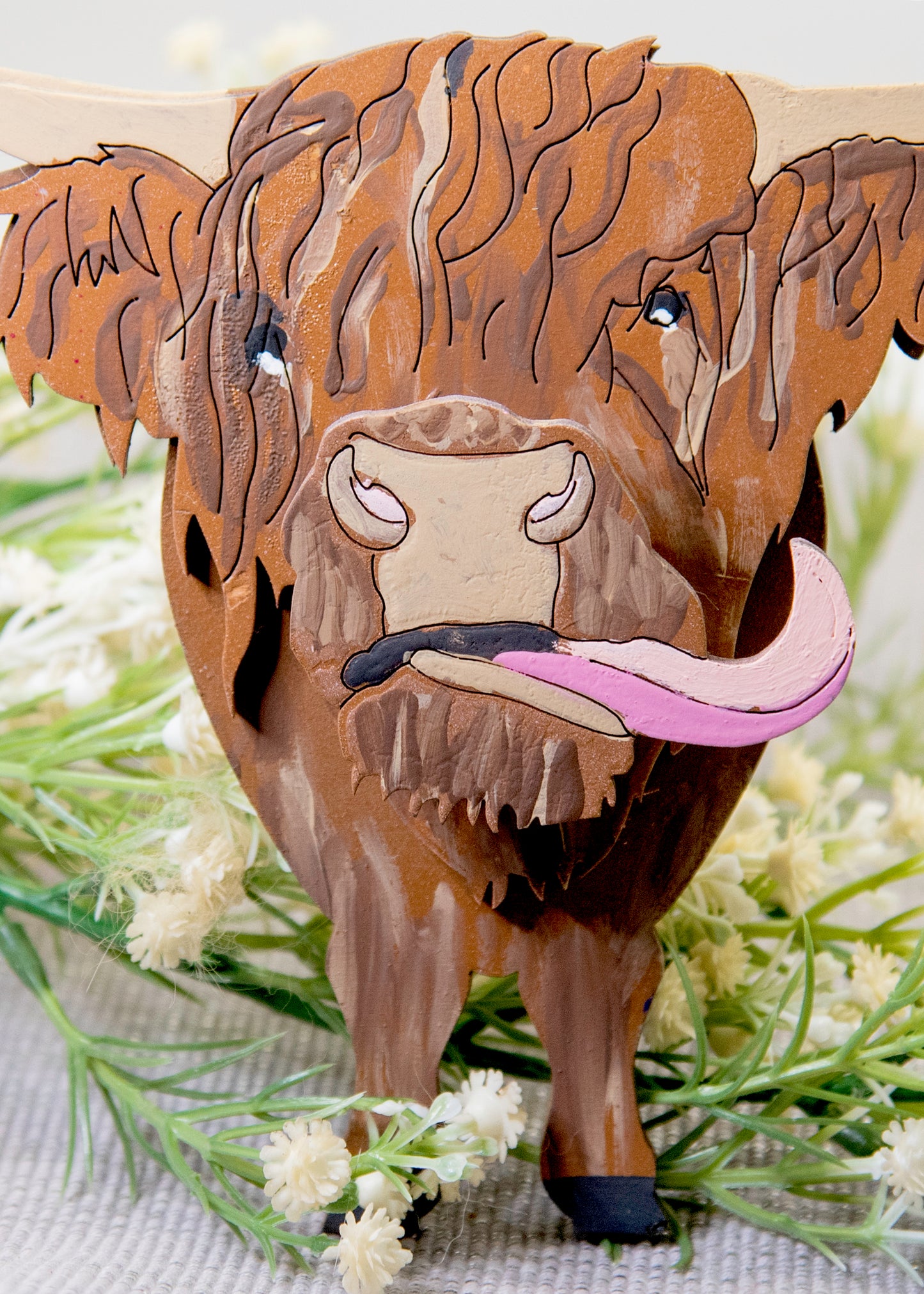 Wooden Highland Cow glasses holder with layered fur detail and curled horns, wearing eyeglasses and sticking out its tongue, surrounded by flowers and a decorative butterfly for a rustic and playful look.
