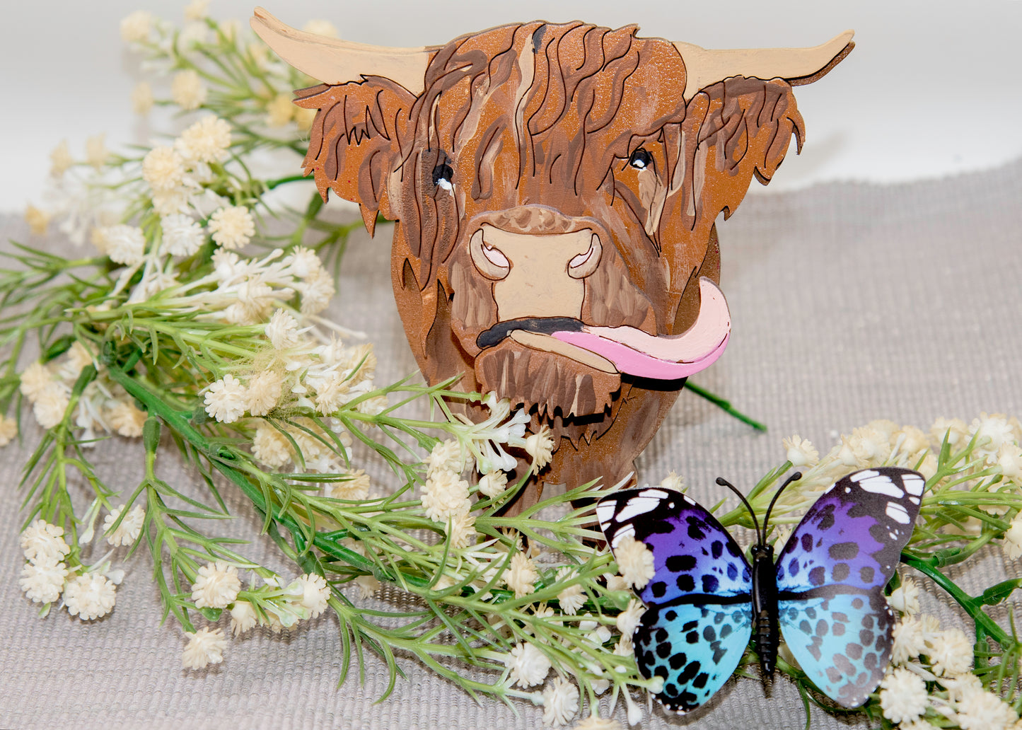 Wooden Highland Cow glasses holder with layered fur detail and curled horns, wearing eyeglasses and sticking out its tongue, surrounded by flowers and a decorative butterfly for a rustic and playful look.
