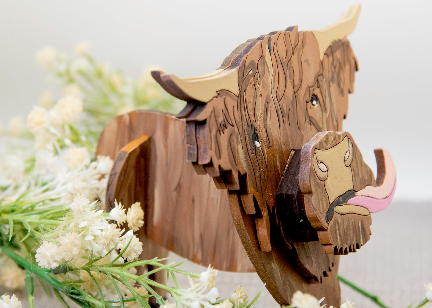 Wooden Highland Cow glasses holder with layered fur detail and curled horns, wearing eyeglasses and sticking out its tongue, surrounded by flowers and a decorative butterfly for a rustic and playful look.