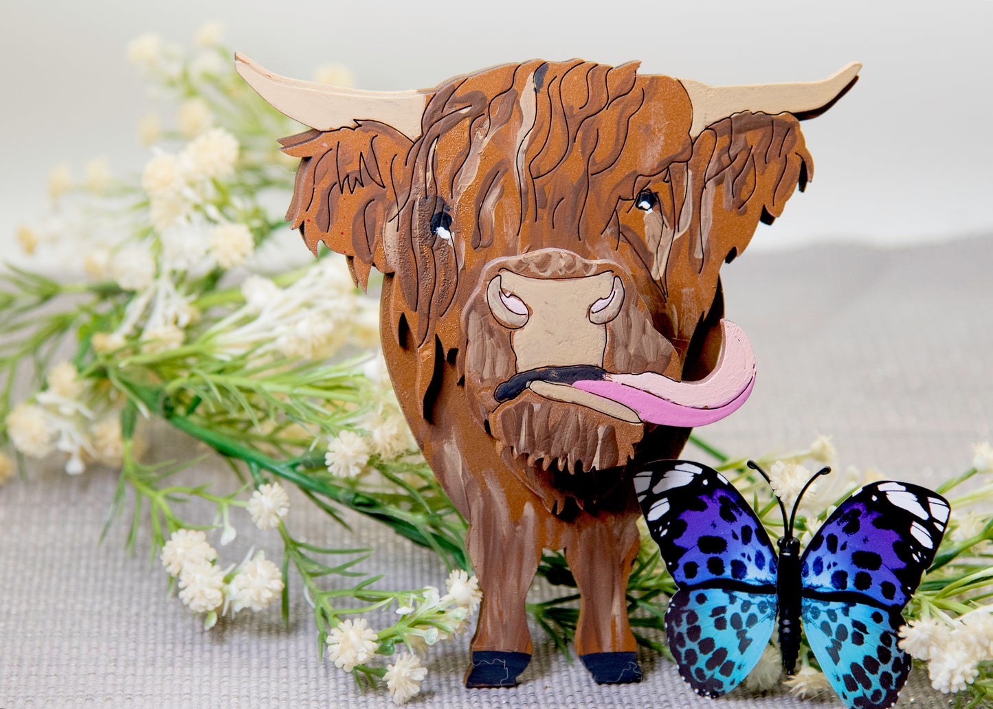 Wooden Highland Cow glasses holder with layered fur detail and curled horns, wearing eyeglasses and sticking out its tongue, surrounded by flowers and a decorative butterfly for a rustic and playful look.