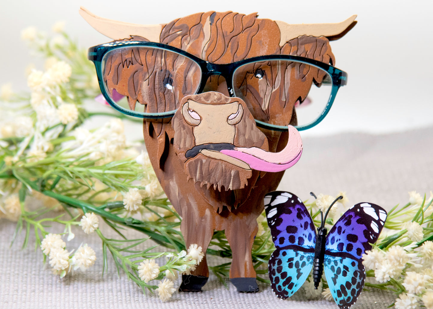 Wooden Highland Cow glasses holder with layered fur detail and curled horns, wearing eyeglasses and sticking out its tongue, surrounded by flowers and a decorative butterfly for a rustic and playful look.