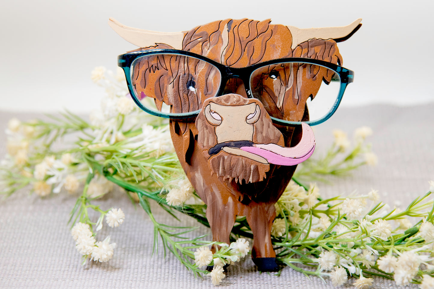 Wooden Highland Cow glasses holder with layered fur detail and curled horns, wearing eyeglasses and sticking out its tongue, surrounded by flowers and a decorative butterfly for a rustic and playful look.