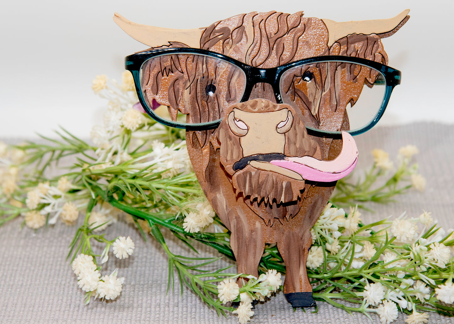 Wooden Highland Cow glasses holder with layered fur detail and curled horns, wearing eyeglasses and sticking out its tongue, surrounded by flowers and a decorative butterfly for a rustic and playful look.