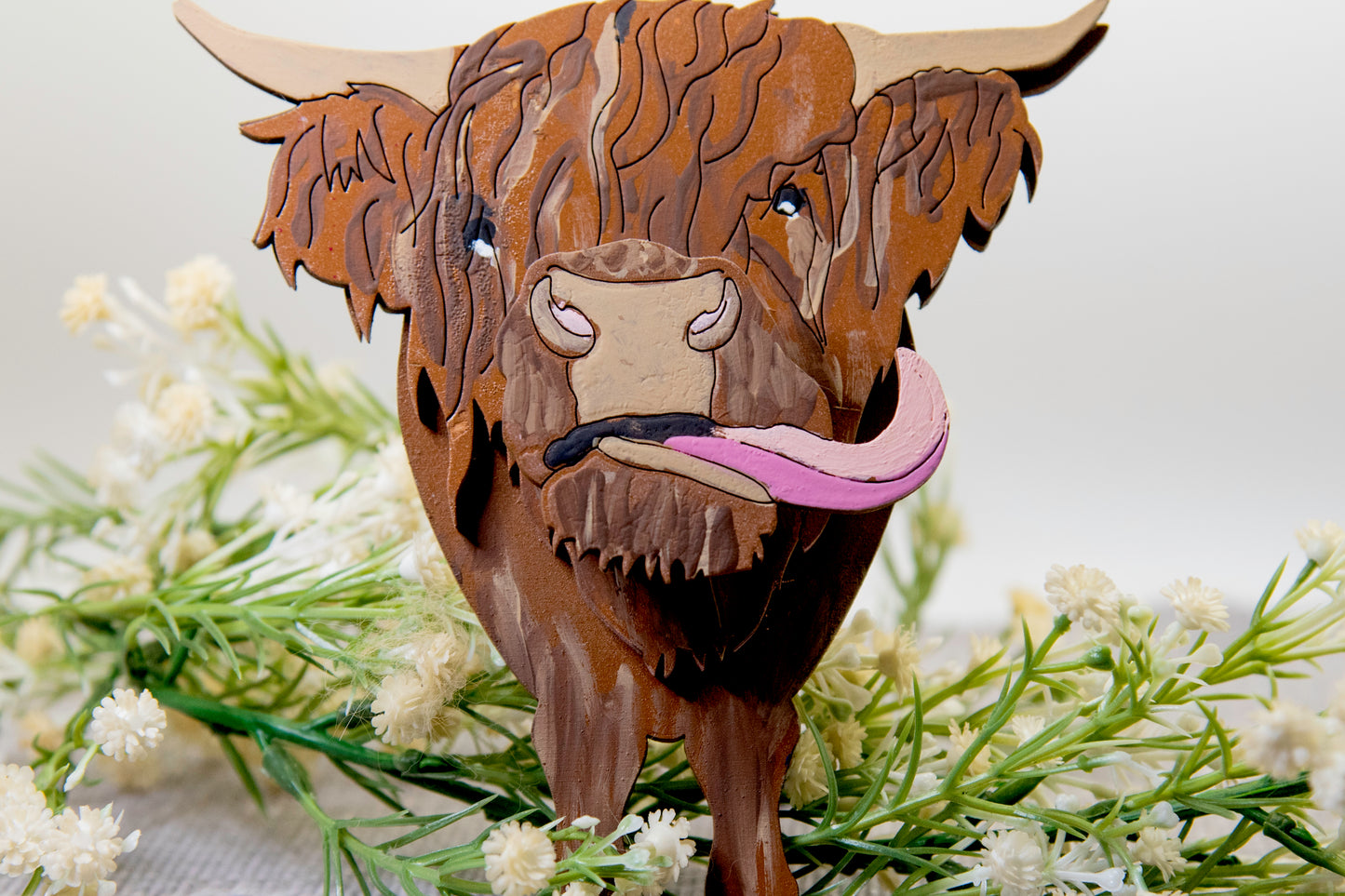 Wooden Highland Cow glasses holder with layered fur detail and curled horns, wearing eyeglasses and sticking out its tongue, surrounded by flowers and a decorative butterfly for a rustic and playful look.