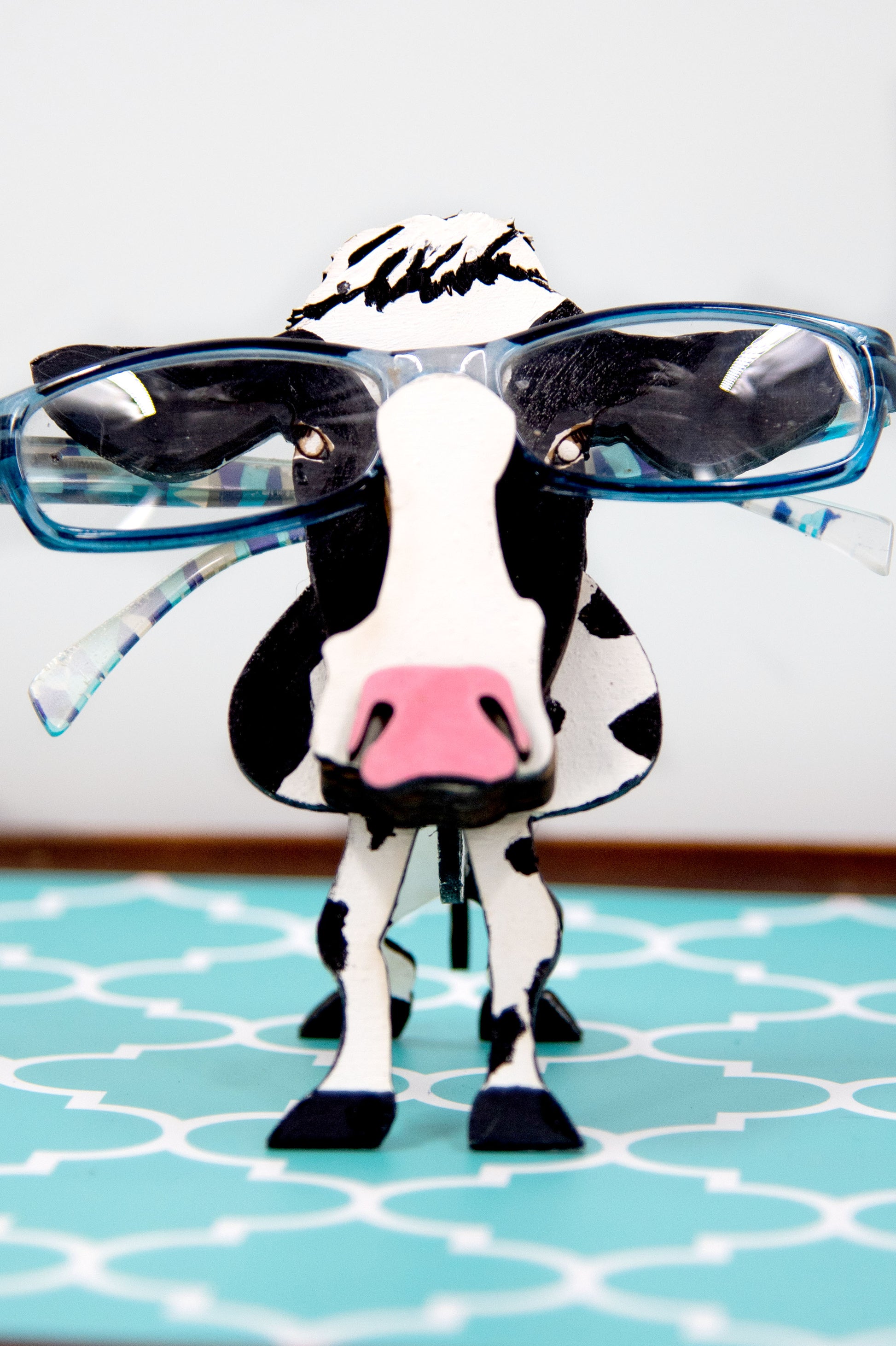 Wooden cow-shaped glasses holder, painted black and white with a layered design, holding a pair of eyeglasses, ideal for farm animal lovers and rustic decor.
