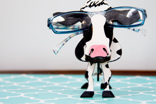 Wooden cow-shaped glasses holder, painted black and white with a layered design, holding a pair of eyeglasses, ideal for farm animal lovers and rustic decor.
