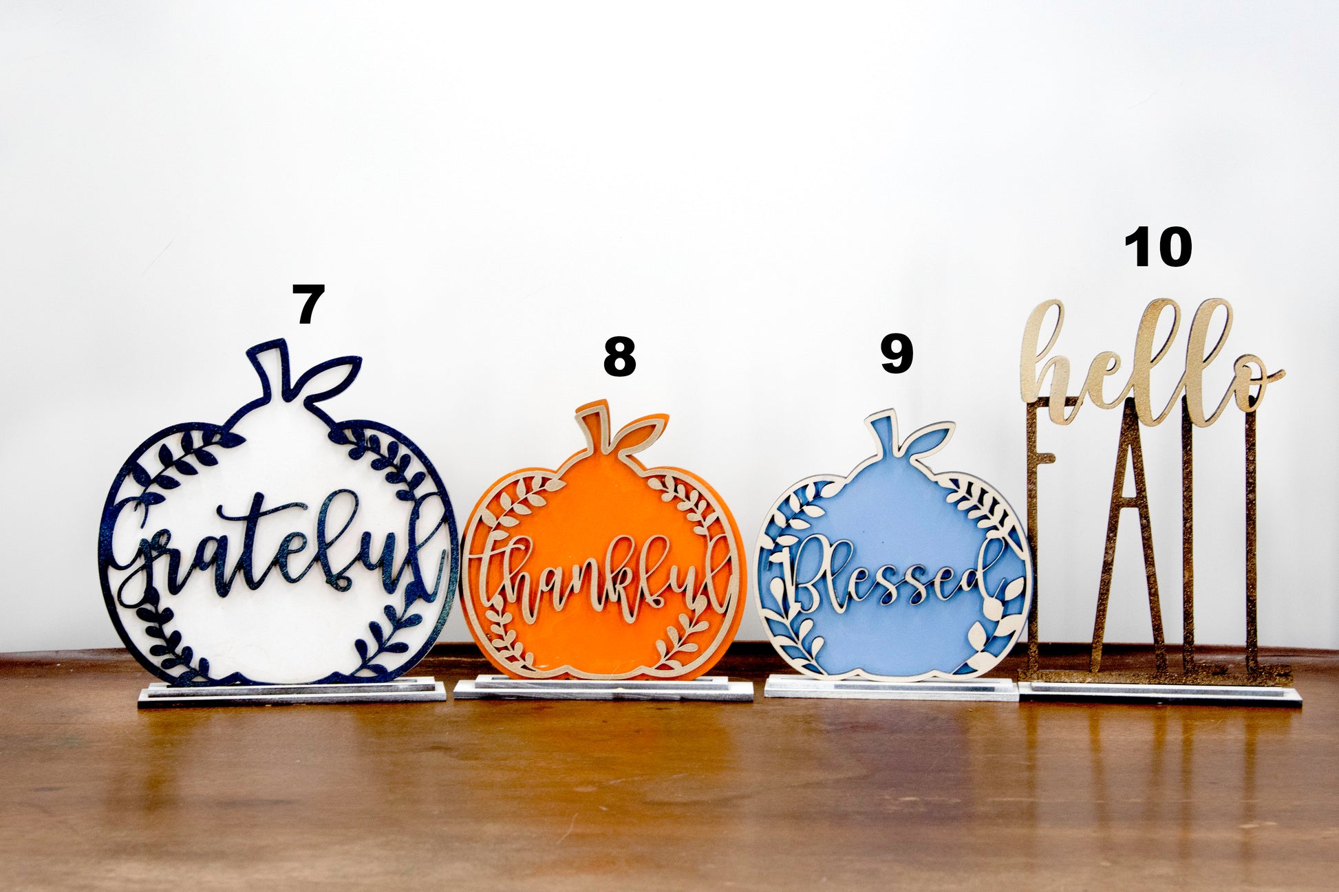Layered standing wooden pumpkins ranging from 5.5 to 8 inches tall, featuring a solid color background and patterned top layer in assorted fall colors and rustic prints. Some designs include engraved words like 'Blessed' and 'Grateful.' Each pumpkin stands on a wooden base, making them ideal for fall shelf decor, Thanksgiving displays, or farmhouse-style seasonal arrangements.
