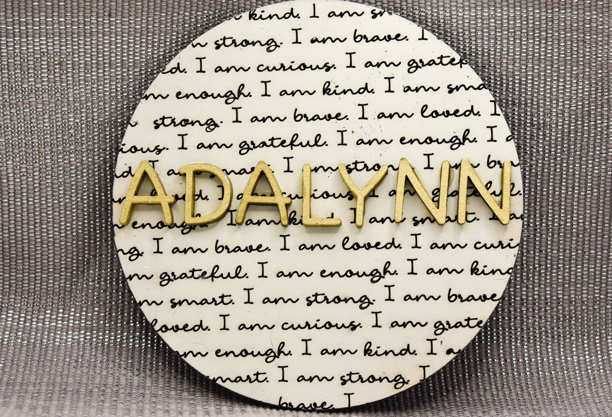 Round wooden name affirmation sign with a custom 3D name on top and engraved positive words like "brave," "kind," and "loved" behind it. Offered in various colors and three sizes, it's perfect for crib name tags, baby announcements, or personalized nursery decor.