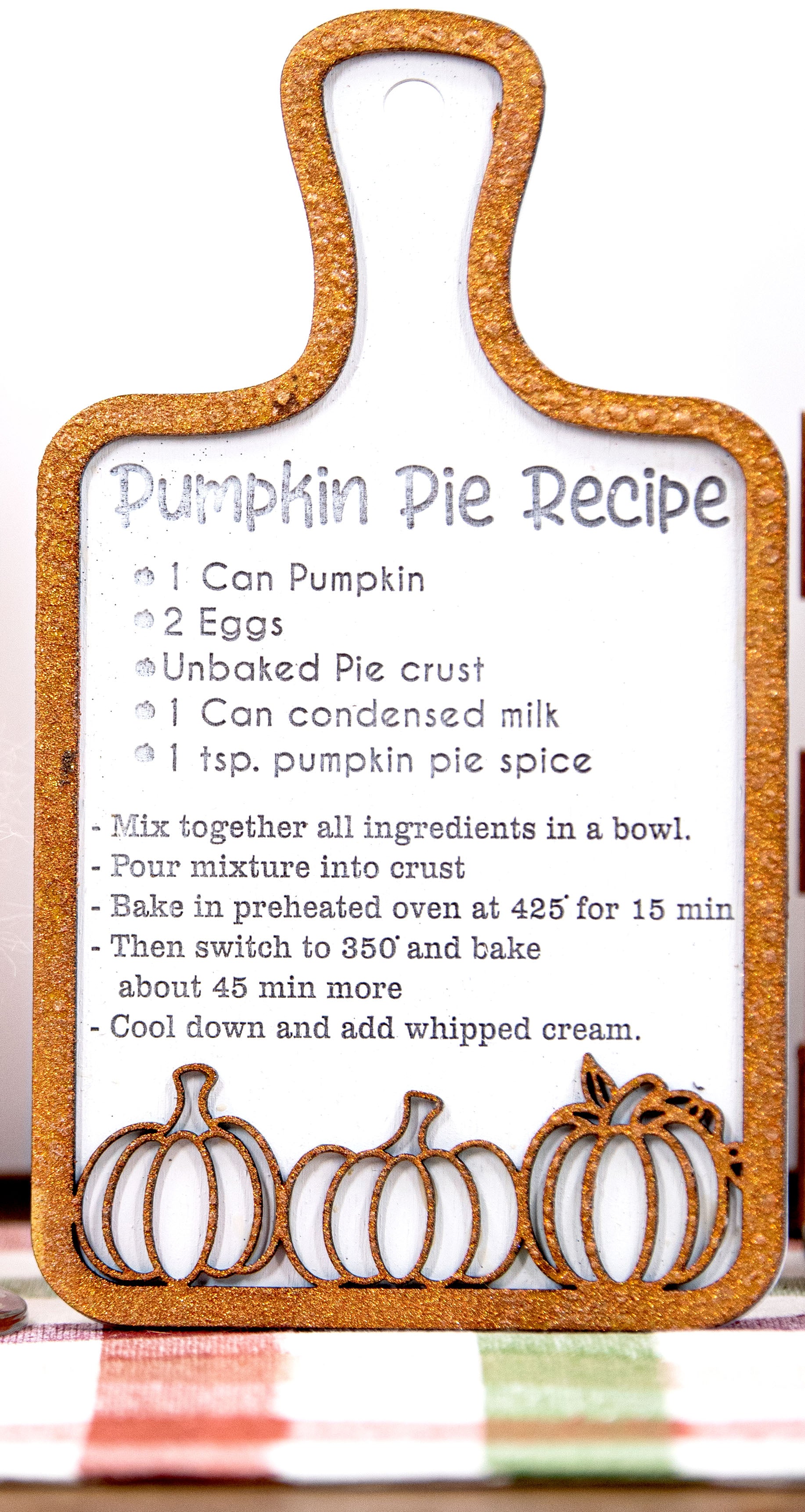 Laser-cut wooden cutting board with engraved pumpkin pie recipe and decorative pumpkins, styled on a fall plaid table runner with pumpkins and wine glass.

