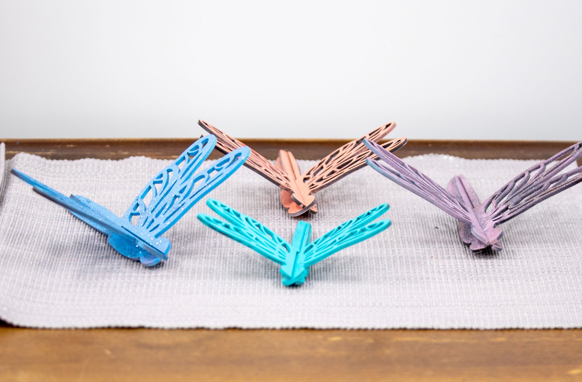 Colorful wooden dragonfly shelf sitters in rustic painted finishes, each shaped with wings and body details, perfect for whimsical spring or summer decor on mantels, shelves, or tiered trays.