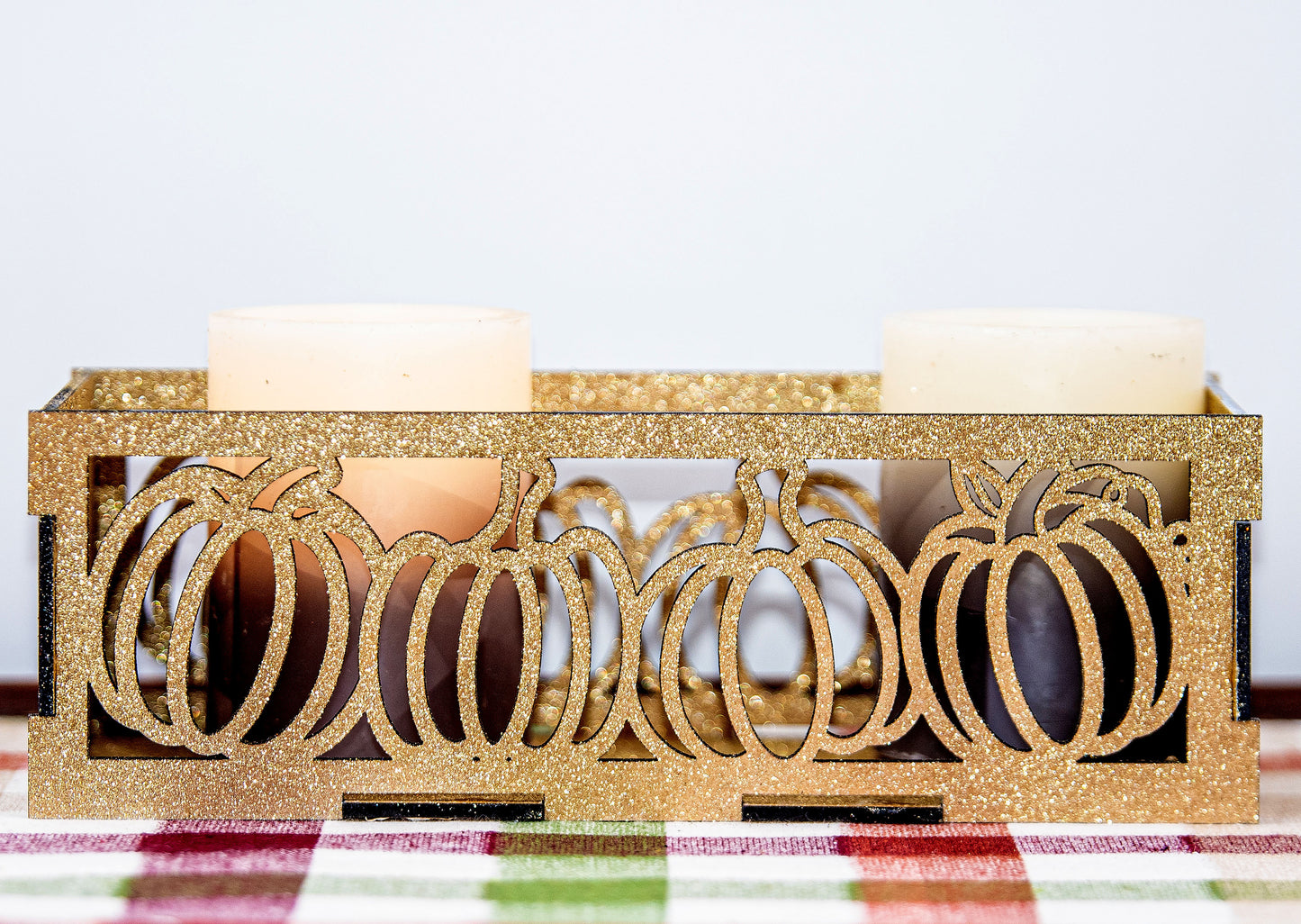 et of three intricately designed metal and wood pumpkin candle holders displayed on a checkered fall table runner. The set includes a tall rectangular lantern, a horizontal centerpiece tray, and a small square holder, each featuring layered pumpkin cutouts that beautifully glow with the warm light of tea candles inside—perfect for cozy fall, Thanksgiving, or farmhouse-style decor.