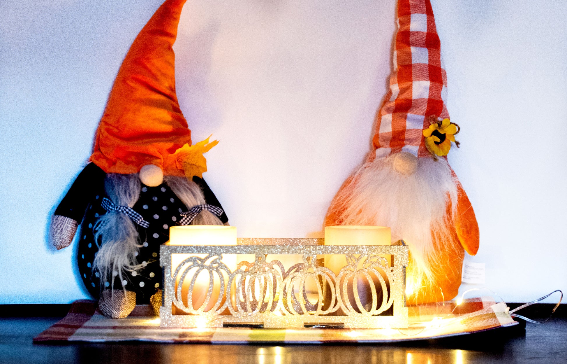 et of three intricately designed metal and wood pumpkin candle holders displayed on a checkered fall table runner. The set includes a tall rectangular lantern, a horizontal centerpiece tray, and a small square holder, each featuring layered pumpkin cutouts that beautifully glow with the warm light of tea candles inside—perfect for cozy fall, Thanksgiving, or farmhouse-style decor.