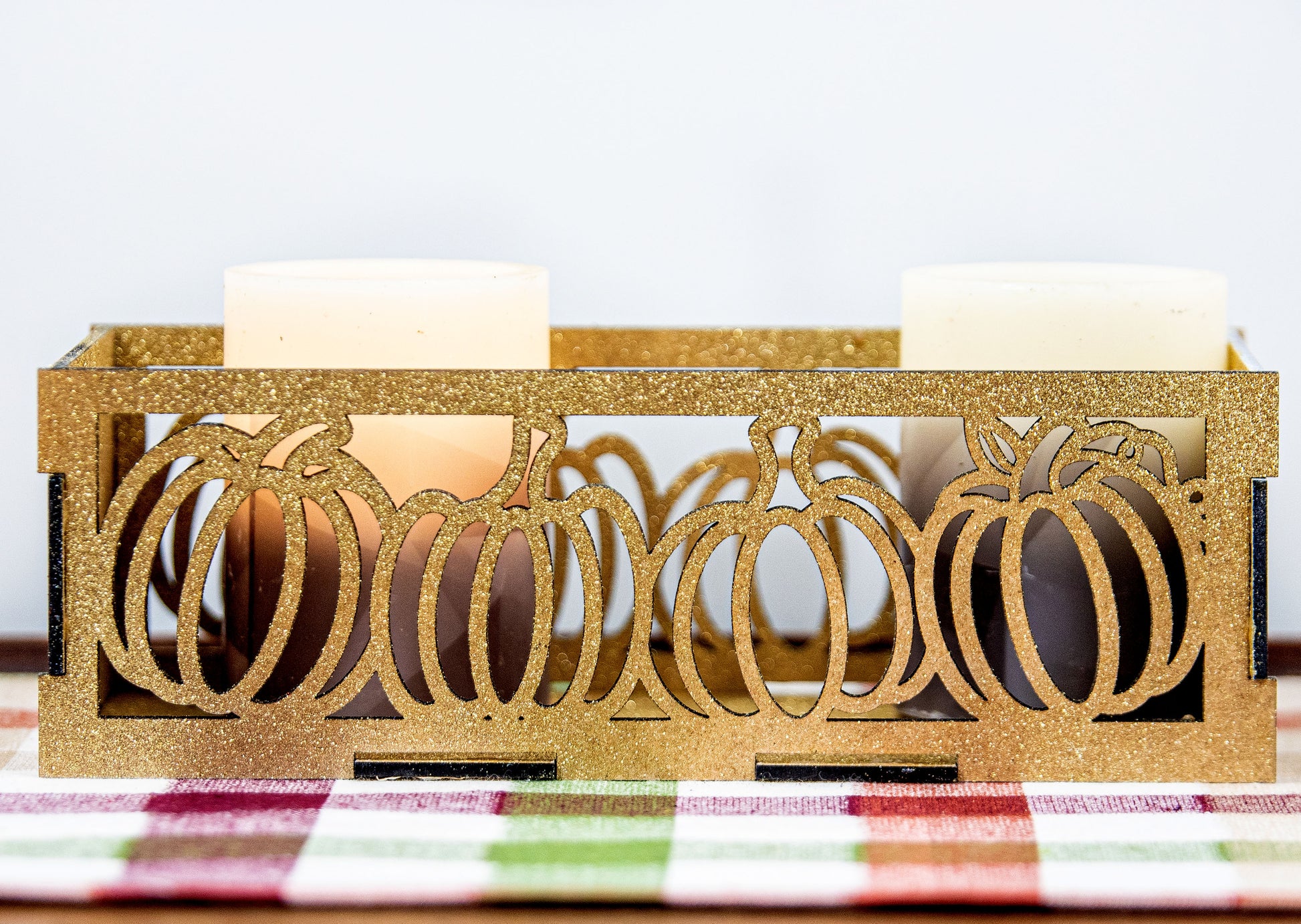 et of three intricately designed metal and wood pumpkin candle holders displayed on a checkered fall table runner. The set includes a tall rectangular lantern, a horizontal centerpiece tray, and a small square holder, each featuring layered pumpkin cutouts that beautifully glow with the warm light of tea candles inside—perfect for cozy fall, Thanksgiving, or farmhouse-style decor.