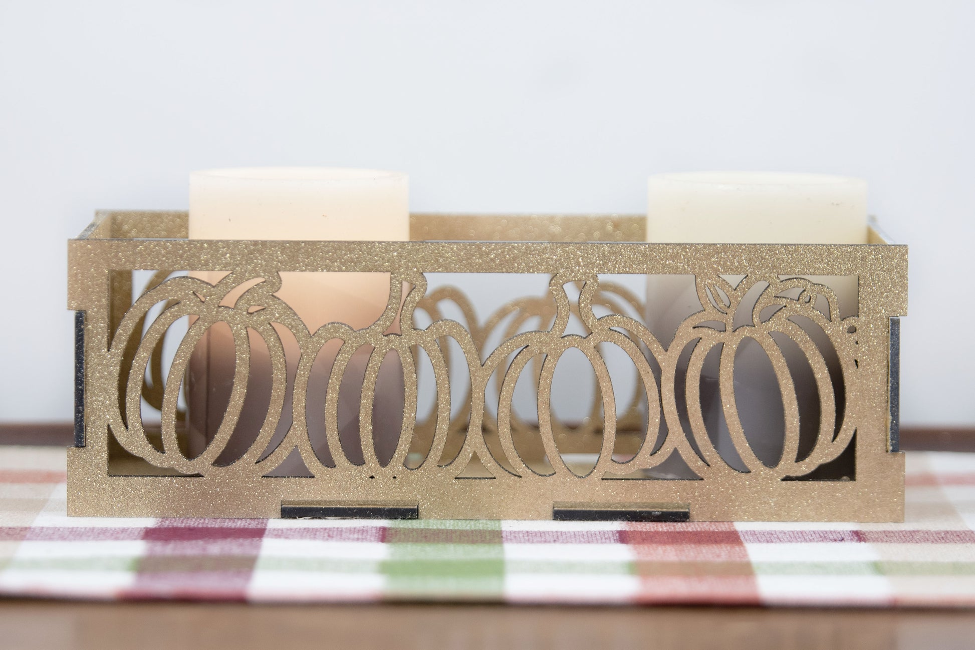 et of three intricately designed metal and wood pumpkin candle holders displayed on a checkered fall table runner. The set includes a tall rectangular lantern, a horizontal centerpiece tray, and a small square holder, each featuring layered pumpkin cutouts that beautifully glow with the warm light of tea candles inside—perfect for cozy fall, Thanksgiving, or farmhouse-style decor.