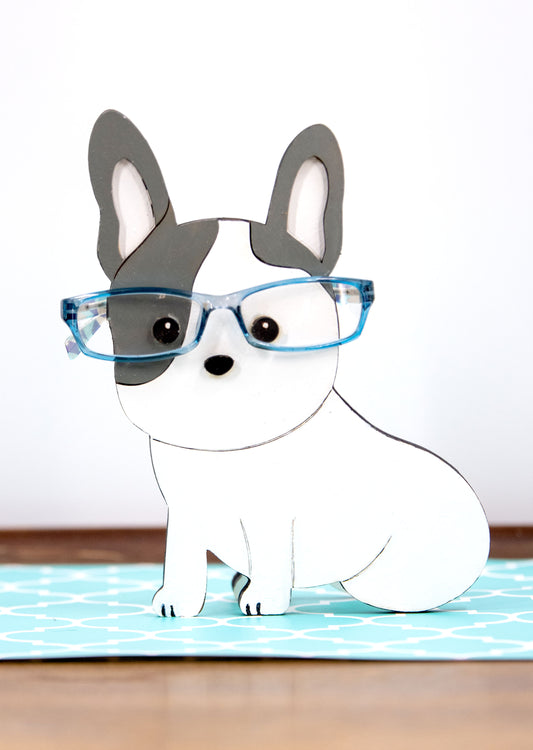 A hand-painted wooden glasses holder shaped like the face of a French Bulldog, featuring detailed ears, muzzle, and facial markings. The stand includes a flat base for stability and is designed to securely hold a pair of eyeglasses on the dog’s nose. Ideal as a decorative desk accessory, nightstand organizer, or pet-themed gift for Frenchie lovers.
