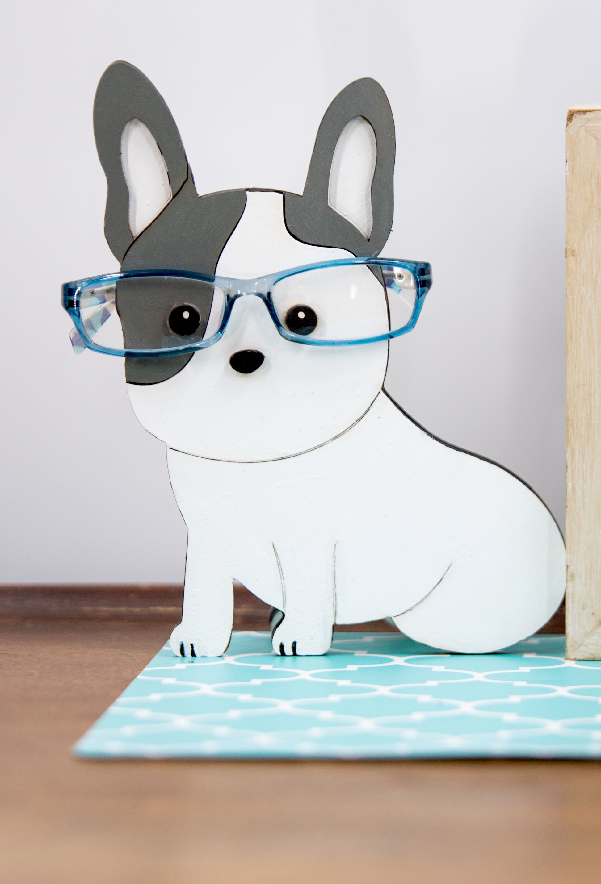 A hand-painted wooden glasses holder shaped like the face of a French Bulldog, featuring detailed ears, muzzle, and facial markings. The stand includes a flat base for stability and is designed to securely hold a pair of eyeglasses on the dog’s nose. Ideal as a decorative desk accessory, nightstand organizer, or pet-themed gift for Frenchie lovers.
