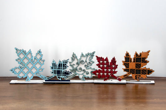 Handcrafted standing wooden leaf decor pieces in layered colors and patterns. Each leaf has a stained background with a cutout patterned top layer, sitting on a flat wood base. Ideal for fall or farmhouse-style decor