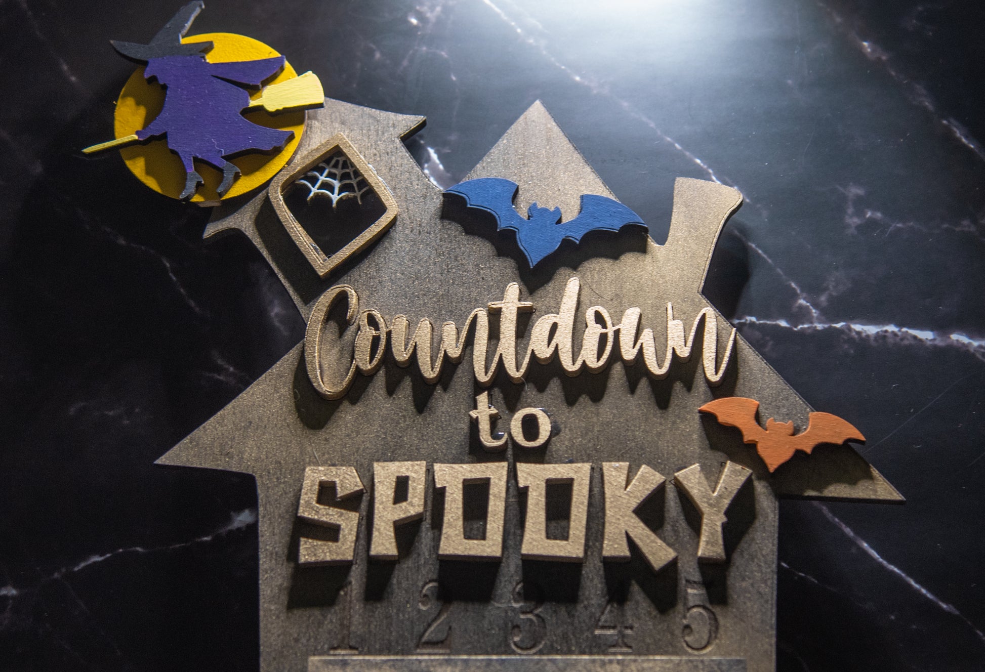 Wooden haunted house-themed Halloween countdown sign with engraved numbers from 1 to 31, painted in classic spooky colors like black, orange, and purple. Features a large haunted mansion design with bats, ghosts, and interchangeable marker icons to track the days until Halloween. Freestanding and available in two sizes, this layered wood sign is perfect for holiday decorating on a mantel, entryway, or shelf display.

