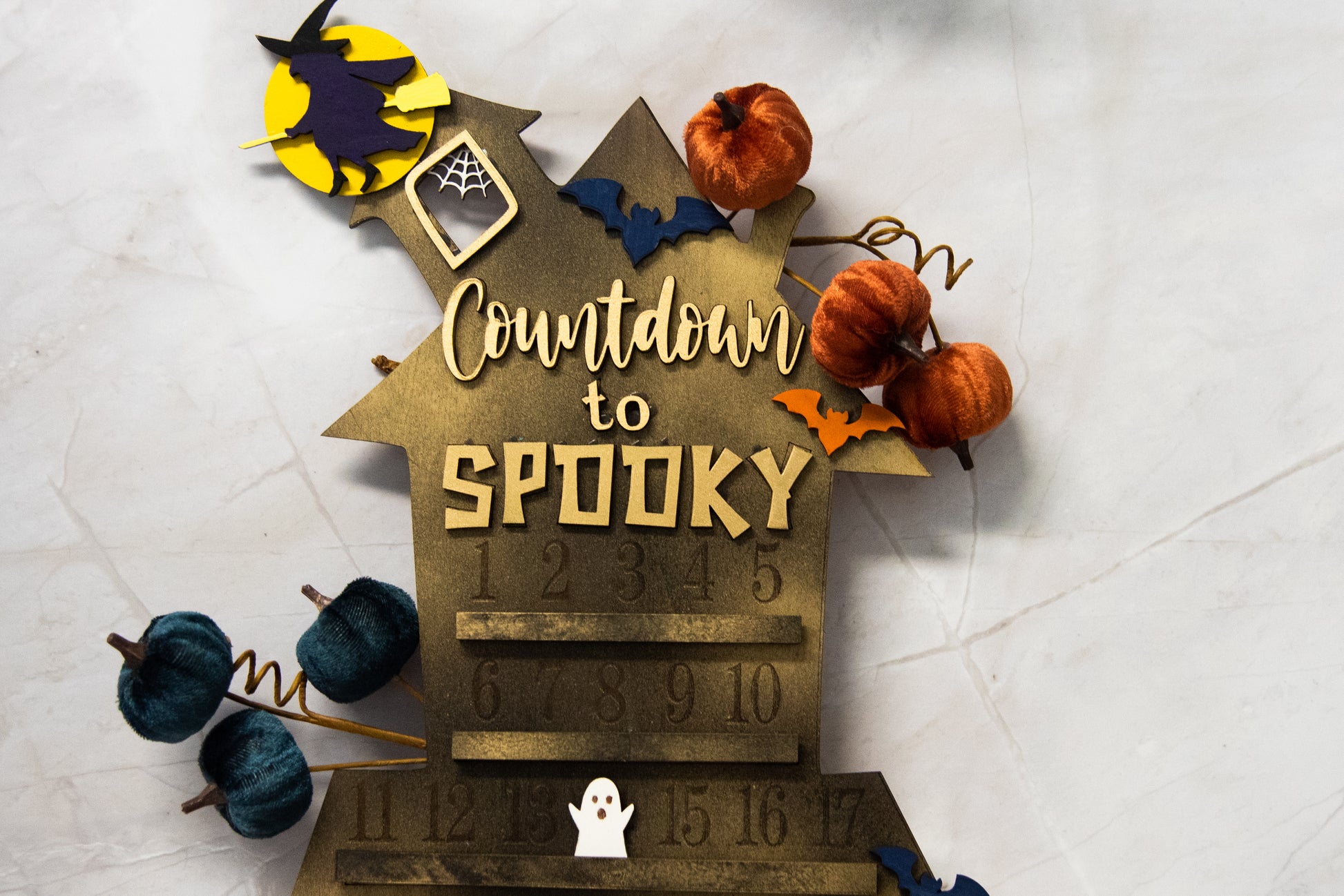 Wooden haunted house-themed Halloween countdown sign with engraved numbers from 1 to 31, painted in classic spooky colors like black, orange, and purple. Features a large haunted mansion design with bats, ghosts, and interchangeable marker icons to track the days until Halloween. Freestanding and available in two sizes, this layered wood sign is perfect for holiday decorating on a mantel, entryway, or shelf display.
