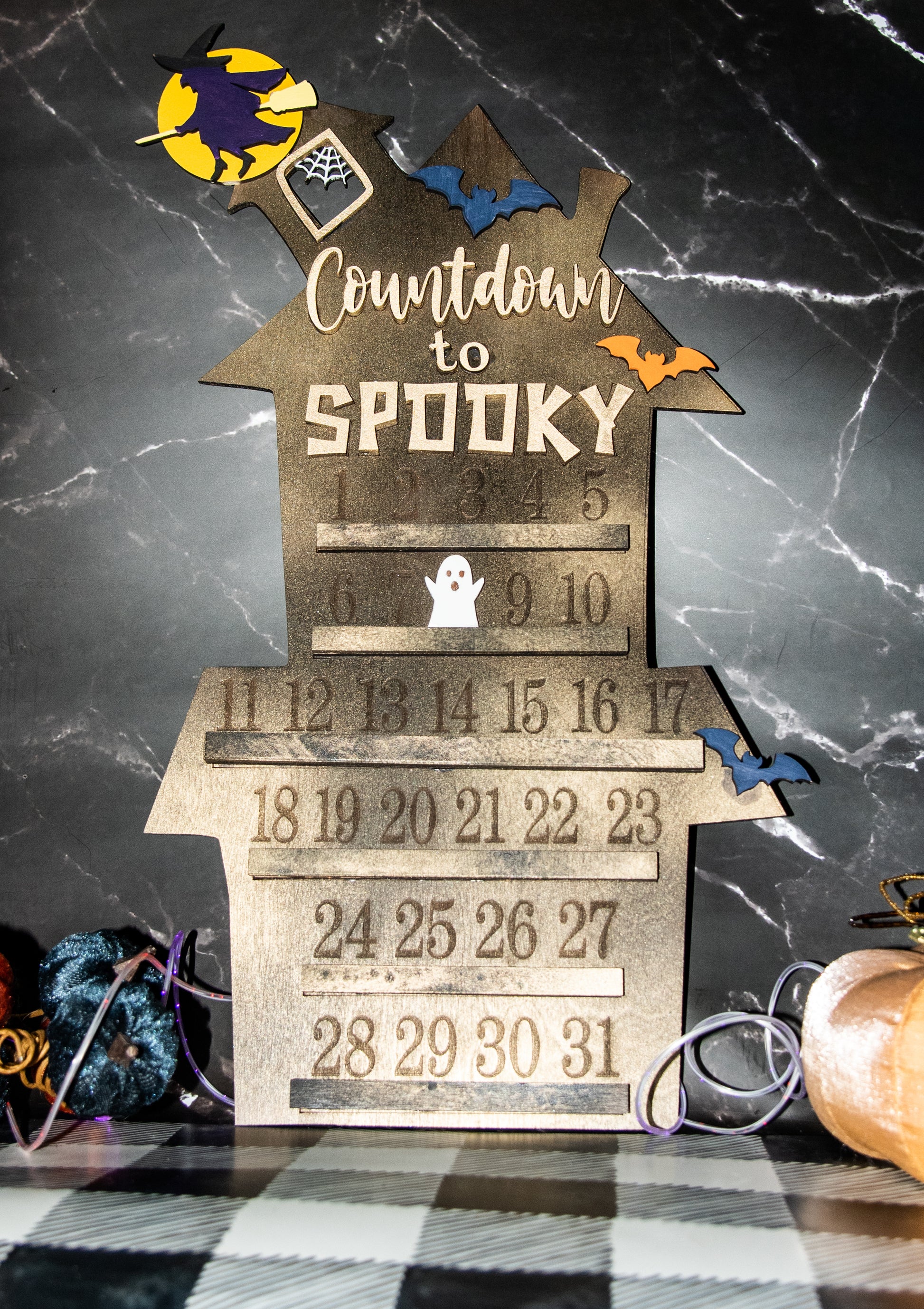 Wooden haunted house-themed Halloween countdown sign with engraved numbers from 1 to 31, painted in classic spooky colors like black, orange, and purple. Features a large haunted mansion design with bats, ghosts, and interchangeable marker icons to track the days until Halloween. Freestanding and available in two sizes, this layered wood sign is perfect for holiday decorating on a mantel, entryway, or shelf display.
