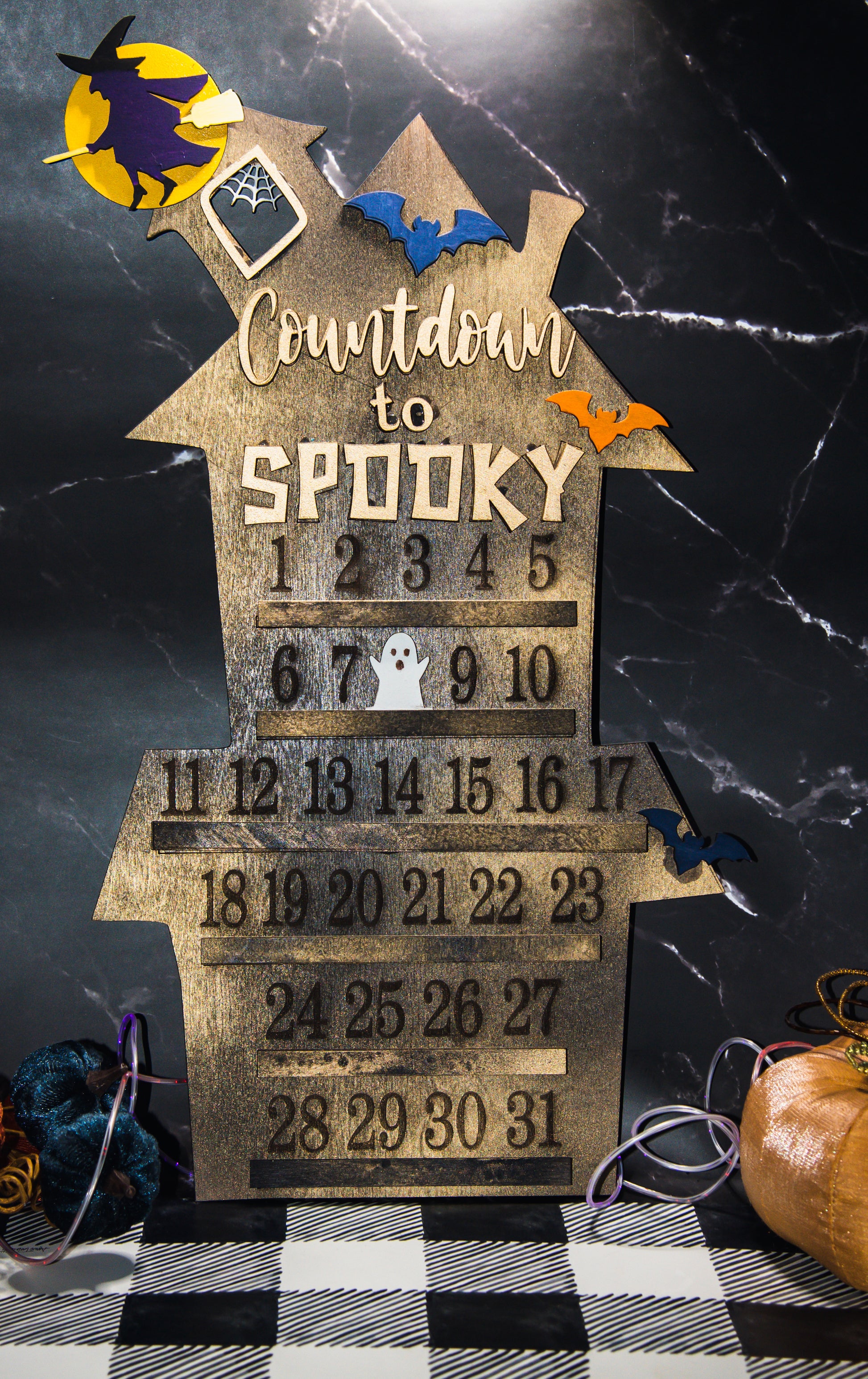 Wooden haunted house-themed Halloween countdown sign with engraved numbers from 1 to 31, painted in classic spooky colors like black, orange, and purple. Features a large haunted mansion design with bats, ghosts, and interchangeable marker icons to track the days until Halloween. Freestanding and available in two sizes, this layered wood sign is perfect for holiday decorating on a mantel, entryway, or shelf display.
